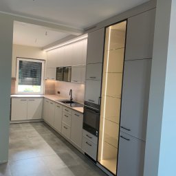 Zakład Stolarsko Budowlany HOLZ Rafał Bembnowicz - Nowoczesna kuchnia z szarymi frontami, czarnymi uchwytami i oświetleniem LED, widok na szafkę z przeszklonym frontem i podświetlonymi półkami.