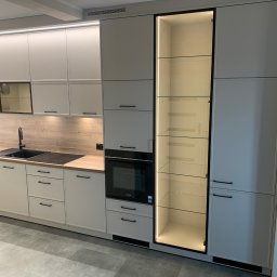 Zakład Stolarsko Budowlany HOLZ Rafał Bembnowicz - Jasna zabudowa kuchenna z beżowymi frontami, czarnymi uchwytami i ramami, drewnianym blatem oraz podświetlaną witryną z półkami ze szkła.