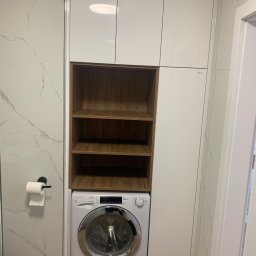 Zakład Stolarsko Budowlany HOLZ Rafał Bembnowicz - Biała zabudowa meblowa w łazience z pralką i drewnianymi półkami wnękowymi, widoczny fragment ściany z marmurowym wzorem i uchwyt na papier toaletowy.