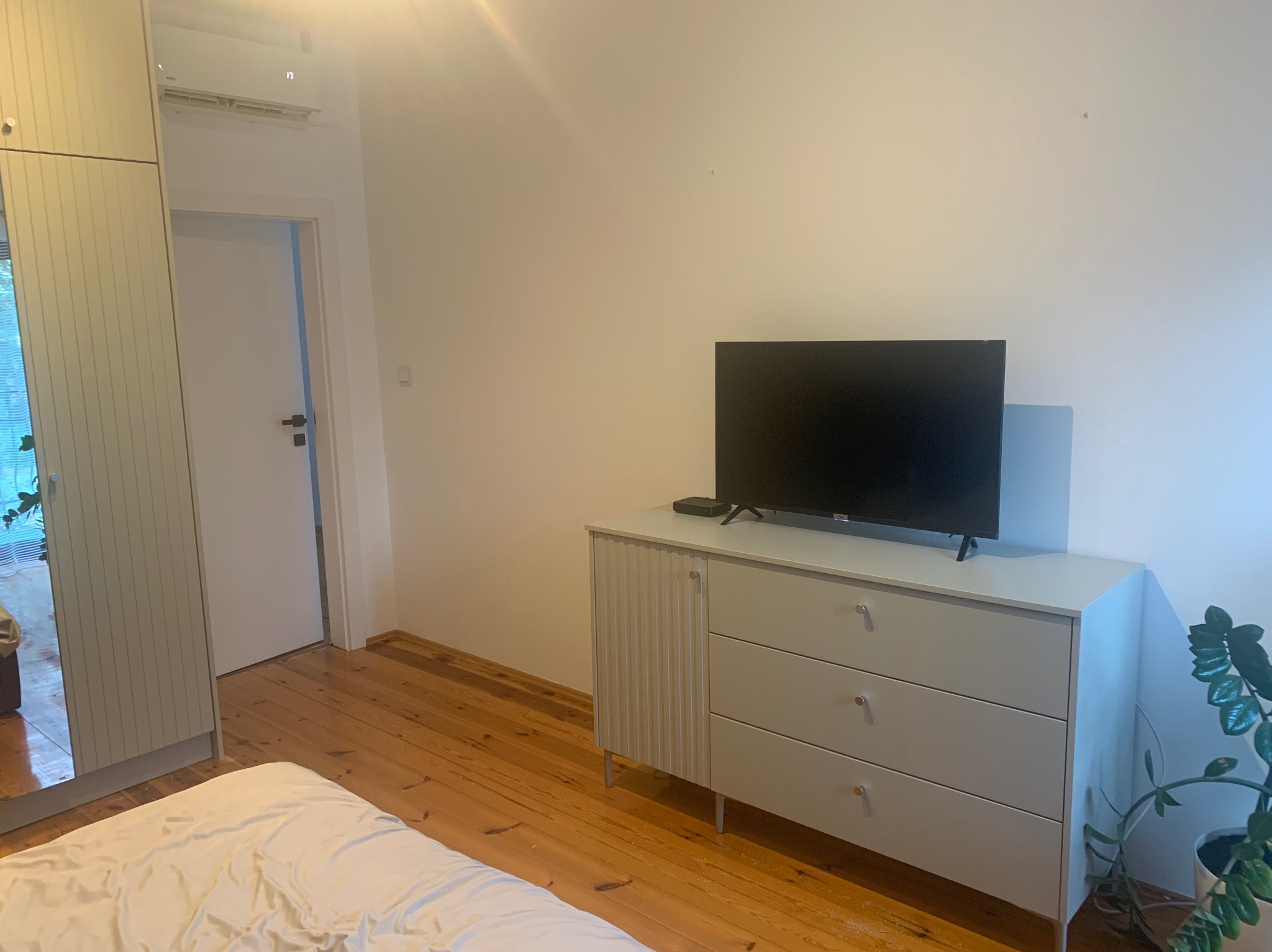 Jasna szafa z trzema szufladami i ryflowanymi drzwiczkami, służąca jako podstawa pod telewizor w sypialni z drewnianą podłogą i widoczną garderobą z lustrzanymi drzwiami.