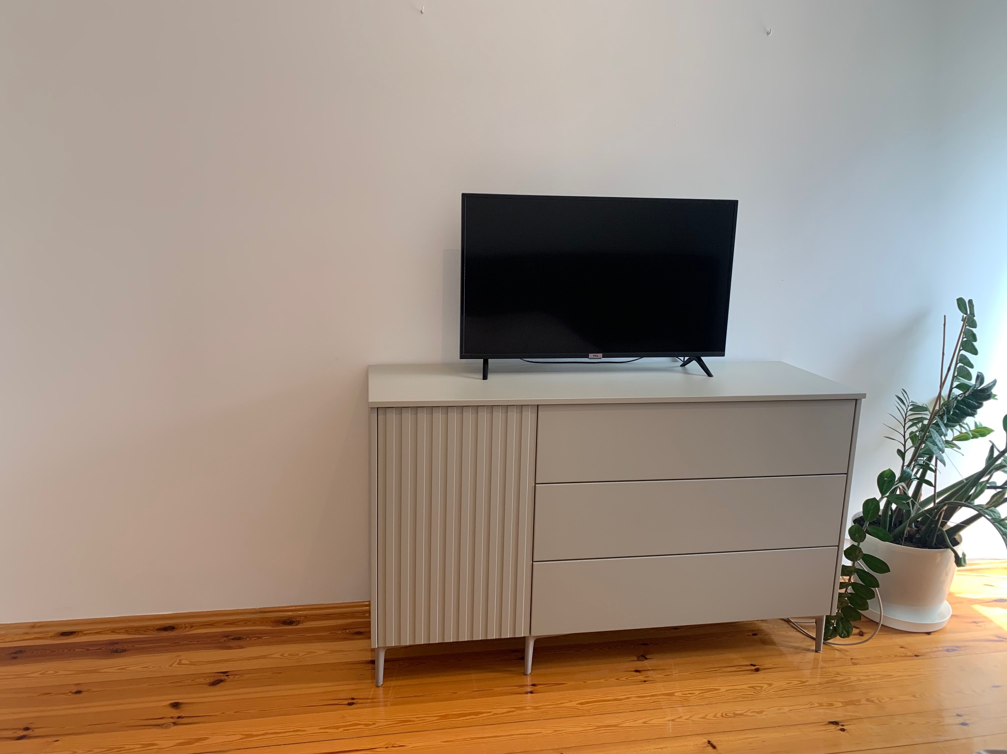 Nowoczesna, szara komoda z trzema szufladami i ryflowanymi drzwiczkami, na metalowych nóżkach, z telewizorem na blacie i rośliną doniczkową obok, na tle białej ściany i drewnianej podłogi.