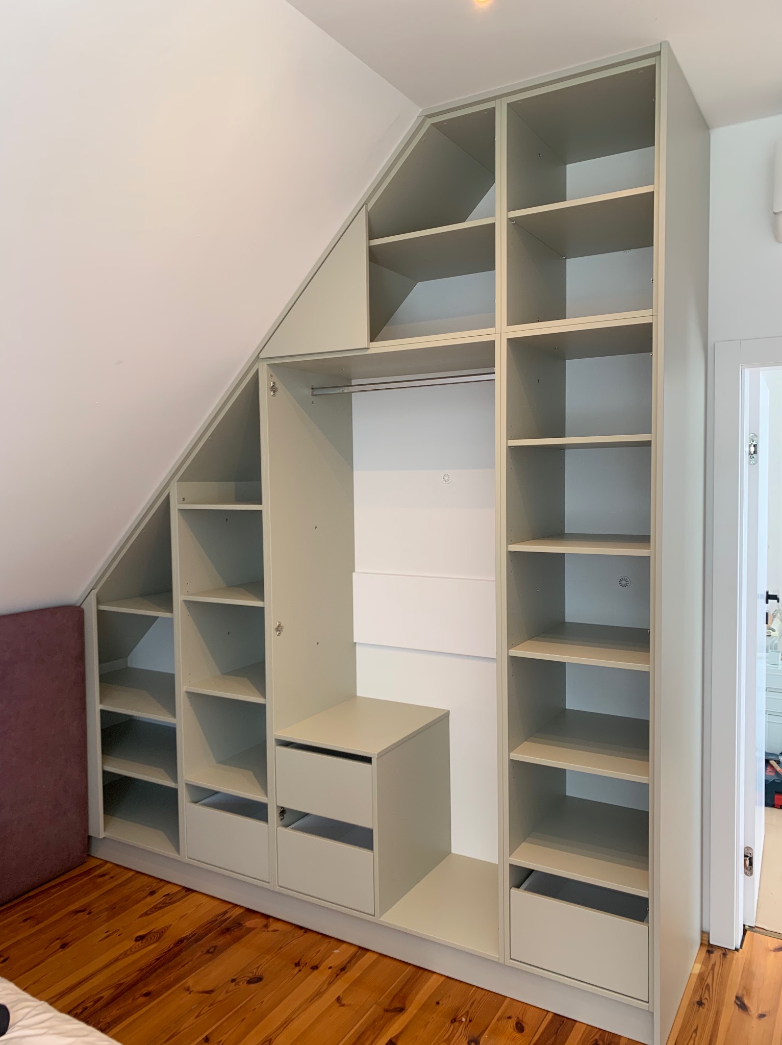 Zabudowa szaf wnękowych w szarym kolorze, dopasowana do skosu poddasza, z otwartymi półkami i szufladami, na drewnianej podłodze.