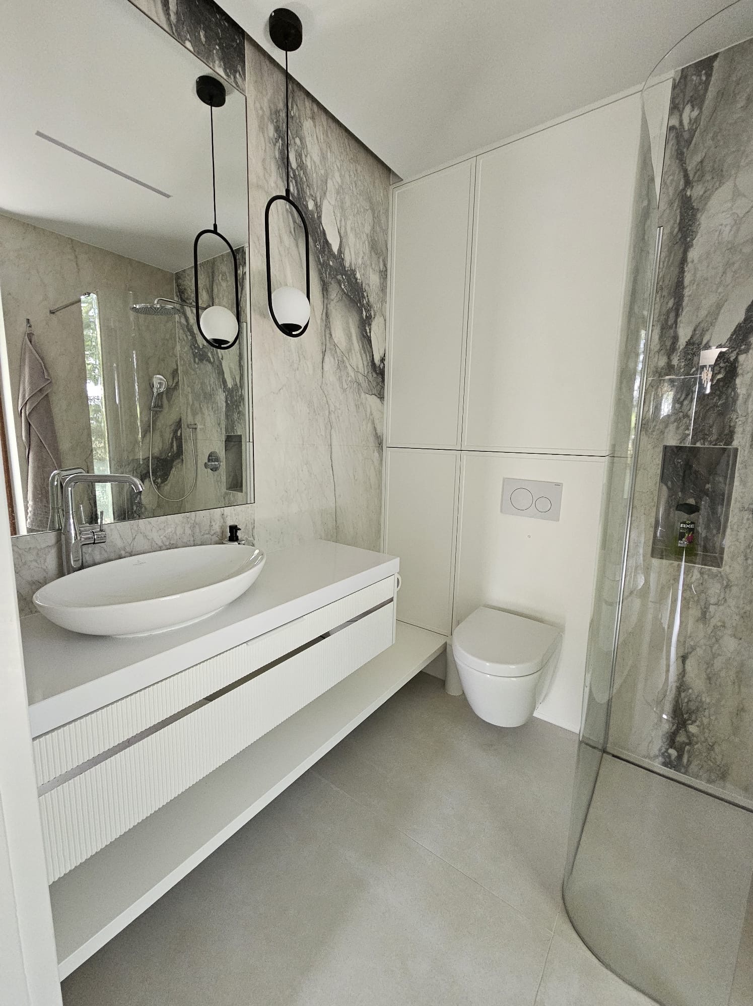 Nowoczesna łazienka z marmurowymi ścianami, białą umywalką nablatową na szafce z frezowanymi frontami, toaletą i kabiną prysznicową ze szklanymi drzwiami.