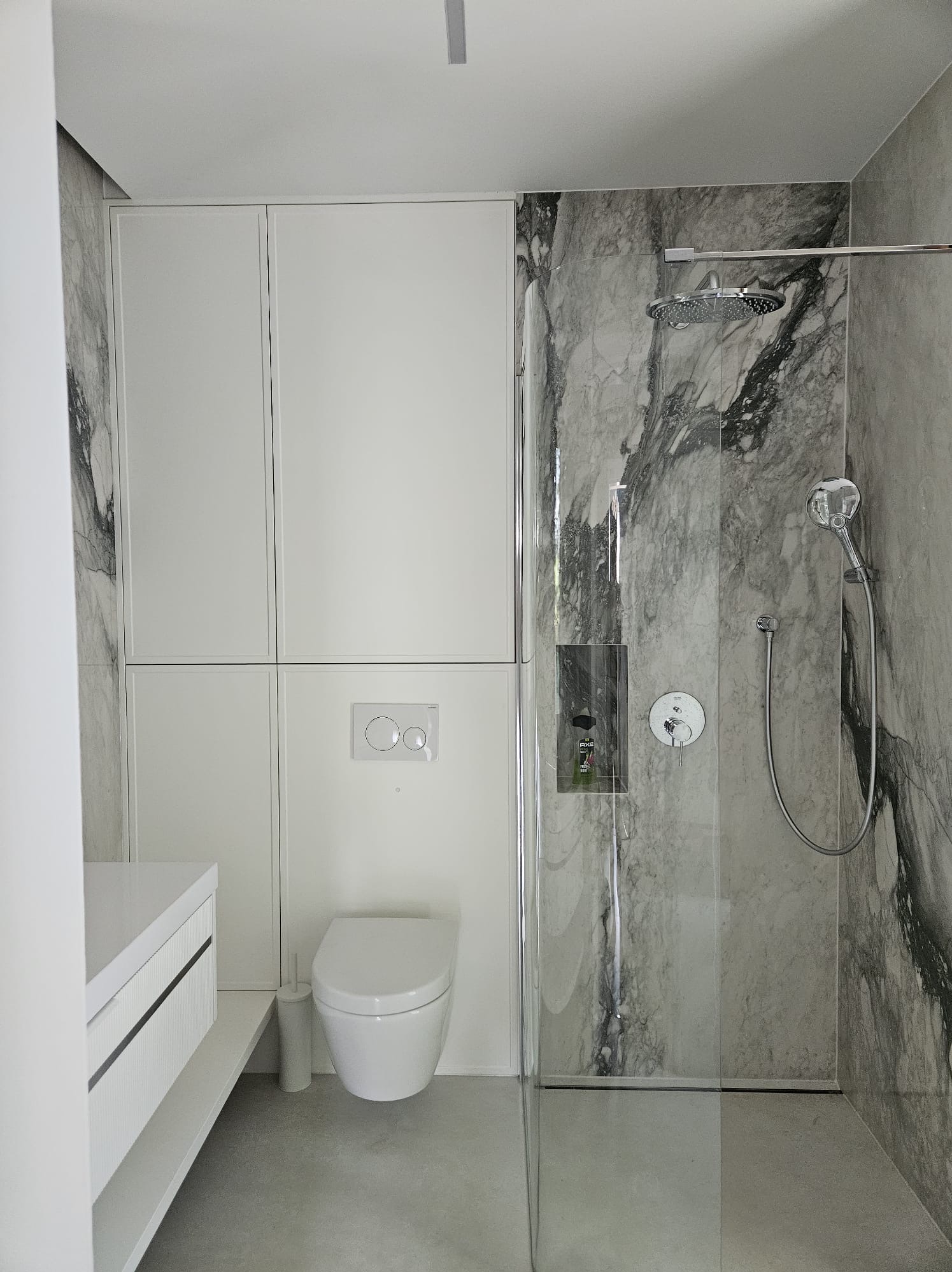 Nowoczesna łazienka z marmurową kabiną prysznicową, białą szafką w zabudowie, podwieszaną toaletą i minimalistyczną umywalką z szafką.