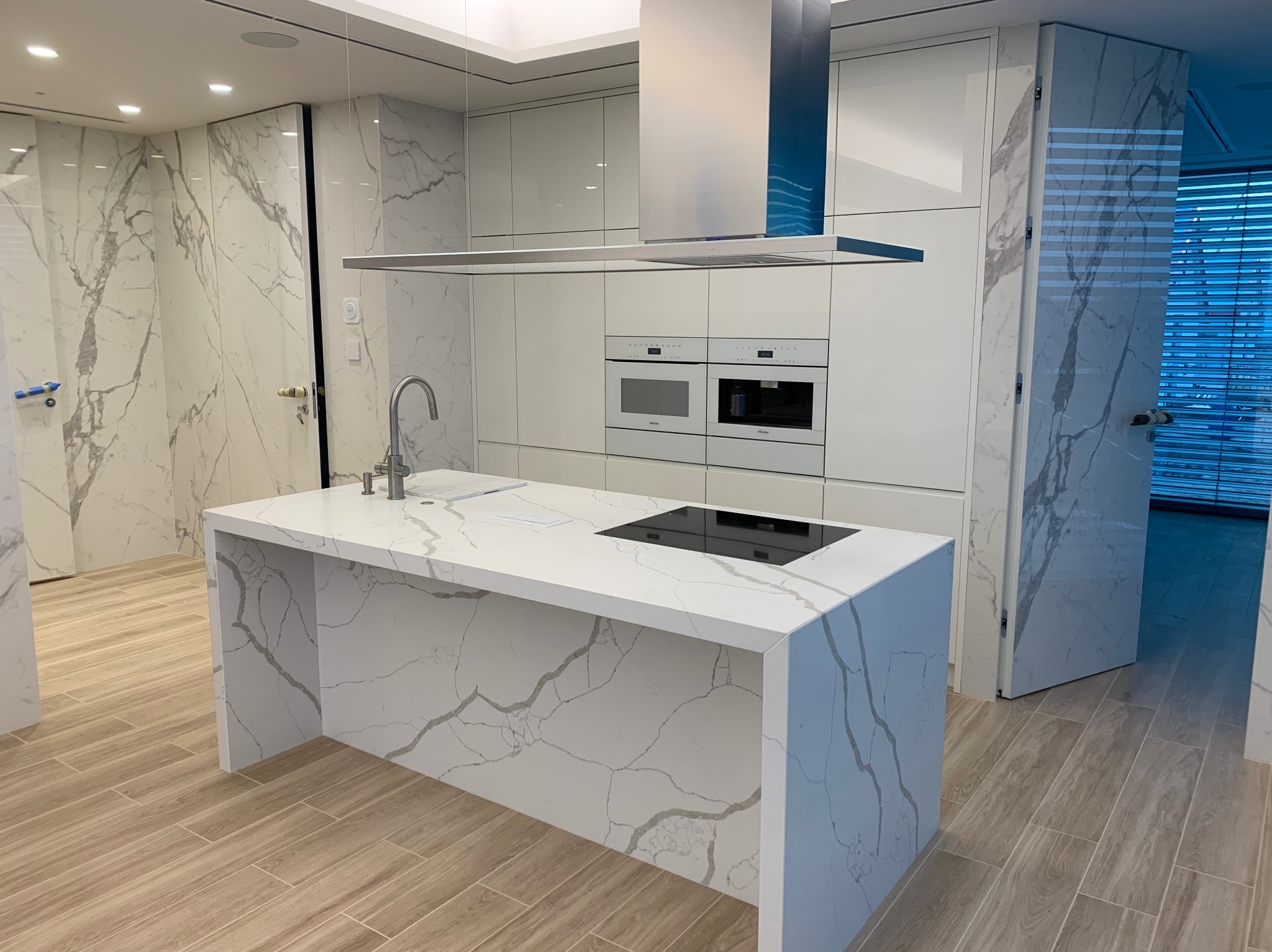 Nowoczesna kuchnia z białymi szafkami i wyspą z blatem imitującym marmur, minimalistyczny okap i zabudowane sprzęty AGD, ściany i drzwi wykończone panelami z wzorem marmuru.