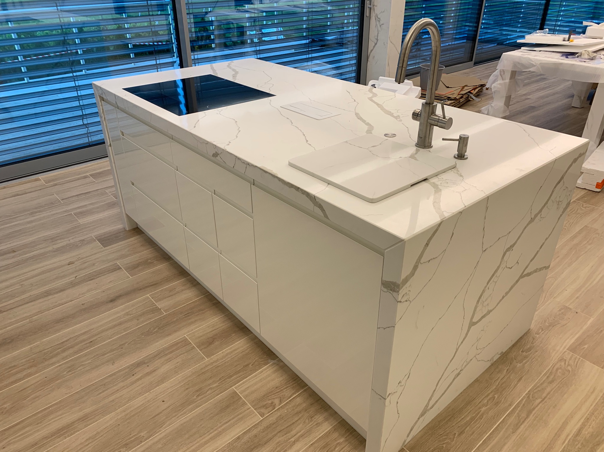 Nowoczesna wyspa kuchenna z białym blatem imitującym marmur, wbudowaną płytą indukcyjną i zlewem ze srebrną baterią, na tle okna z żaluzjami.