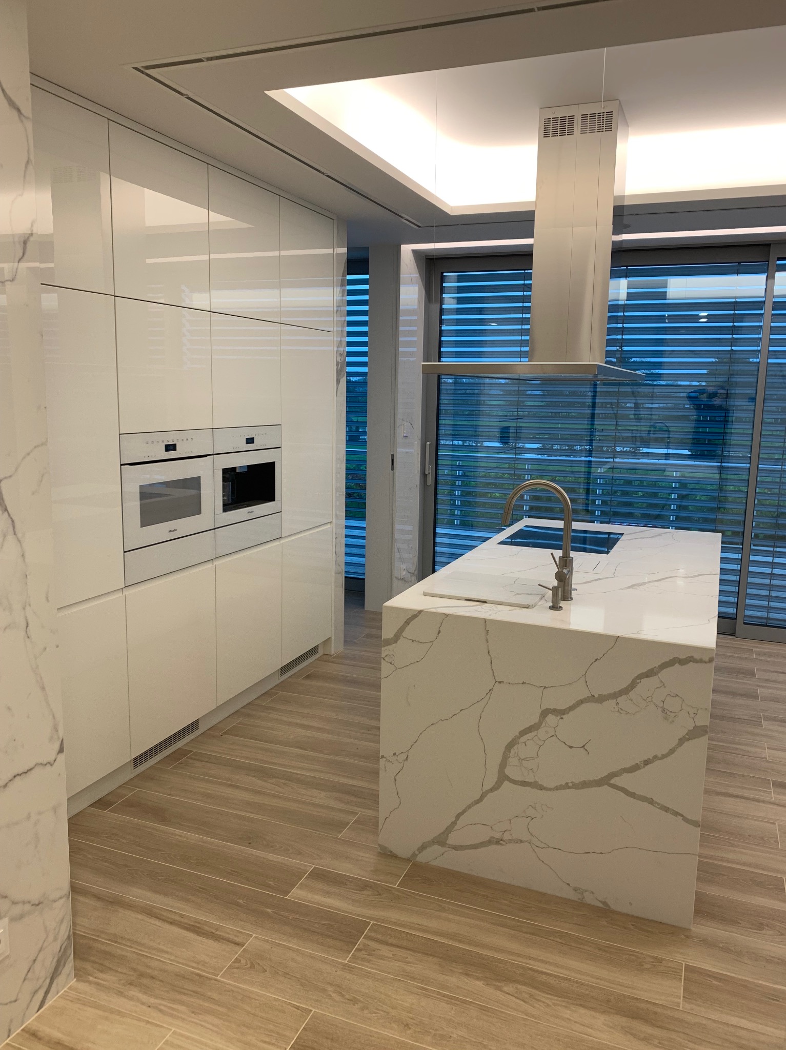 Nowoczesna biała kuchnia z wyspą i marmurowym blatem, oświetlenie LED, zabudowane sprzęty Miele, widok na zewnątrz przez żaluzje.