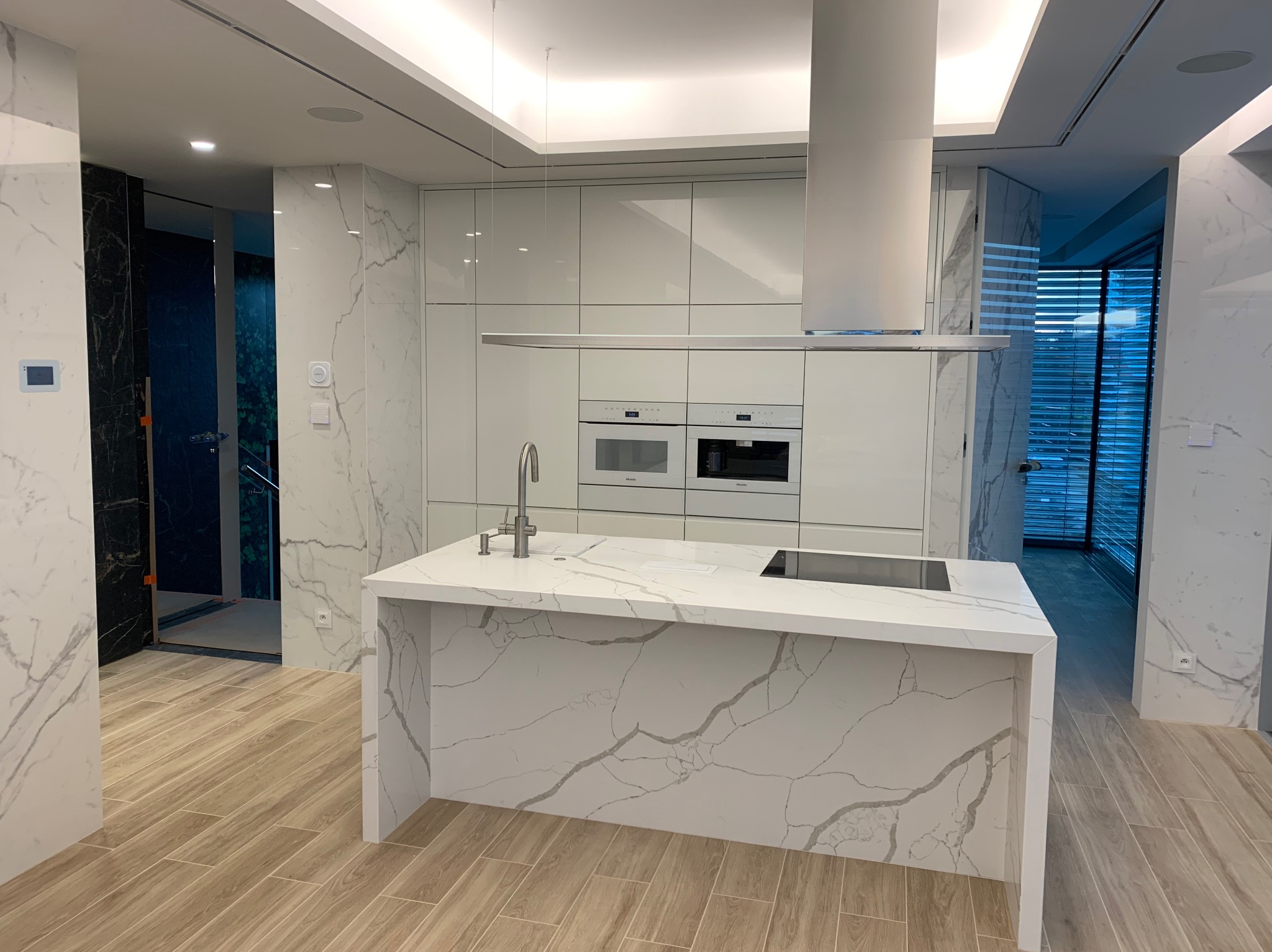 Nowoczesna kuchnia z białą wyspą i marmurowymi akcentami, widok na drzwi z ciemnego drewna w tle.