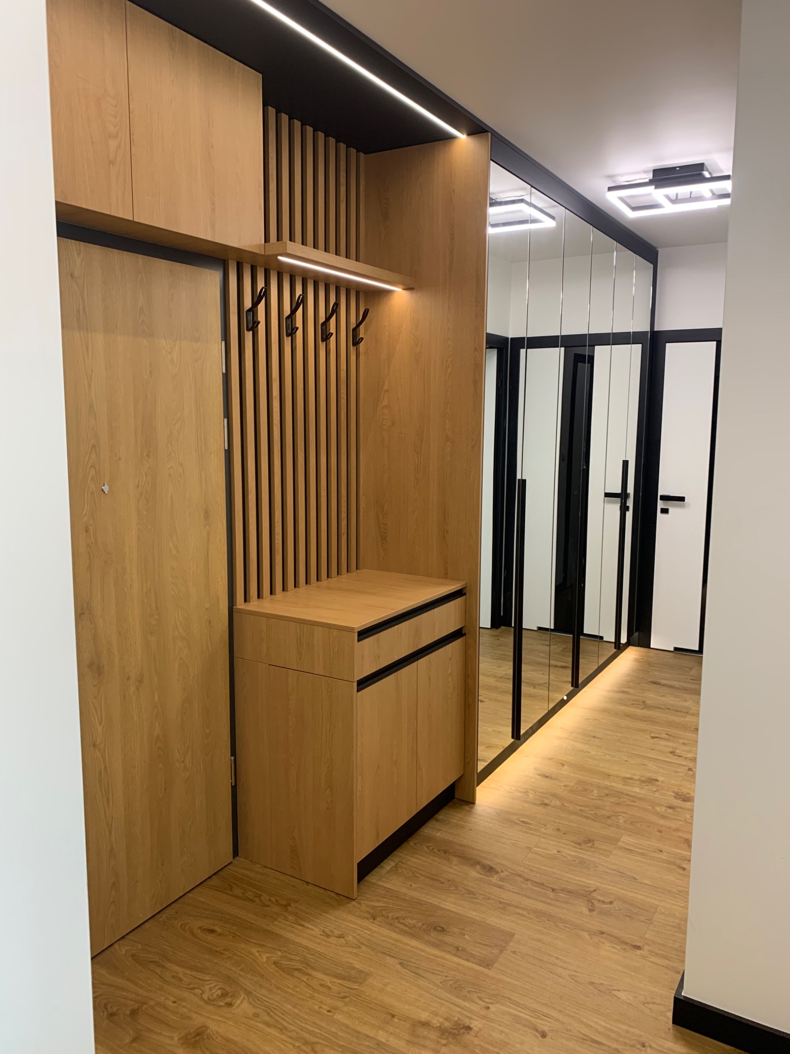 Nowoczesny przedpokój z zabudową z jasnego drewna: szafka na buty z szufladami, panel z wieszakami, szafka wisząca i lustro w wąskich ramach. Całość oświetlona listwami LED.