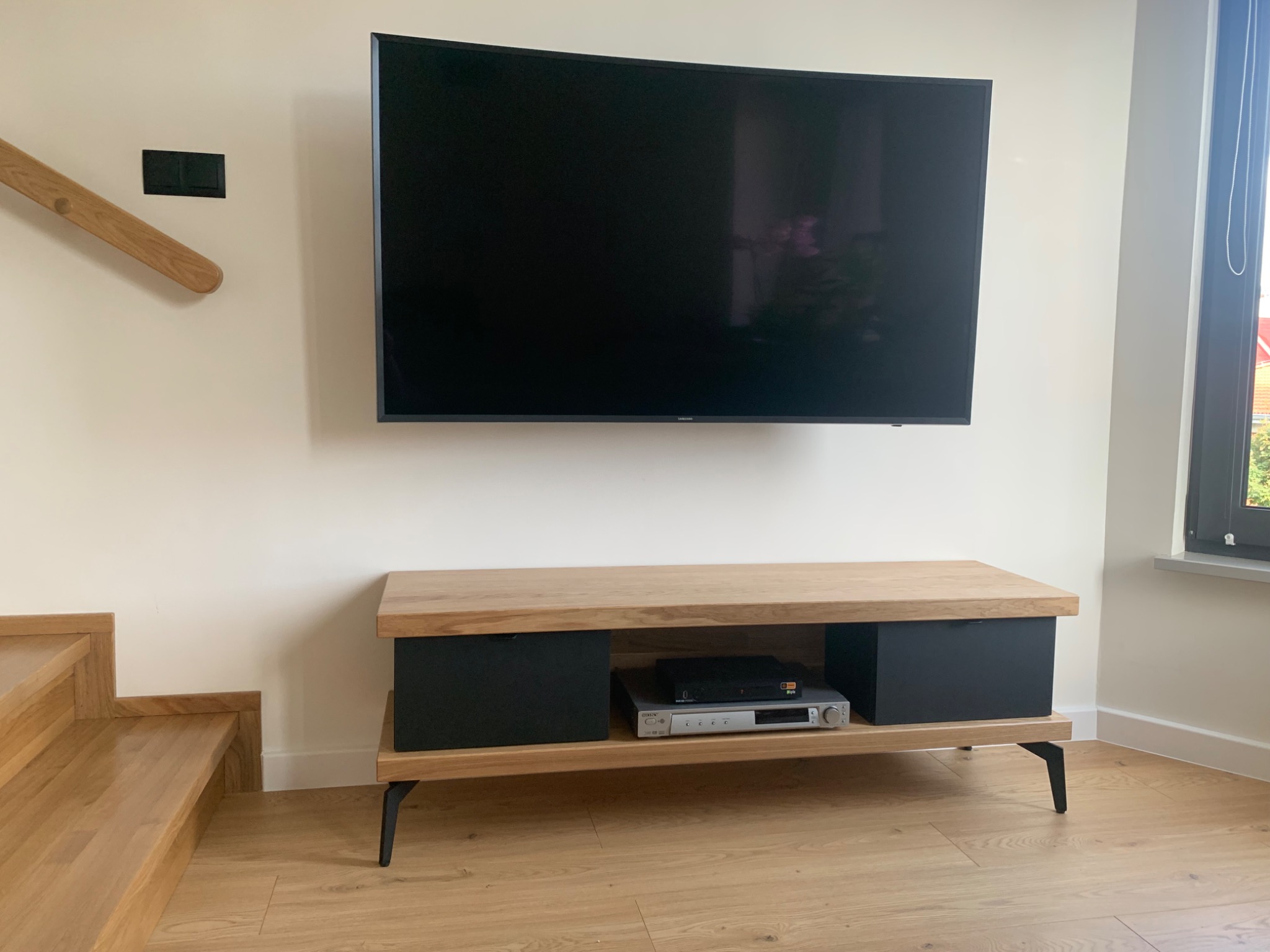 Nowoczesna szafka RTV z drewnianym blatem i czarnymi frontami szuflad, ustawiona pod zawieszonym telewizorem. Widoczne drewniane schody i fragment okna z roletą.