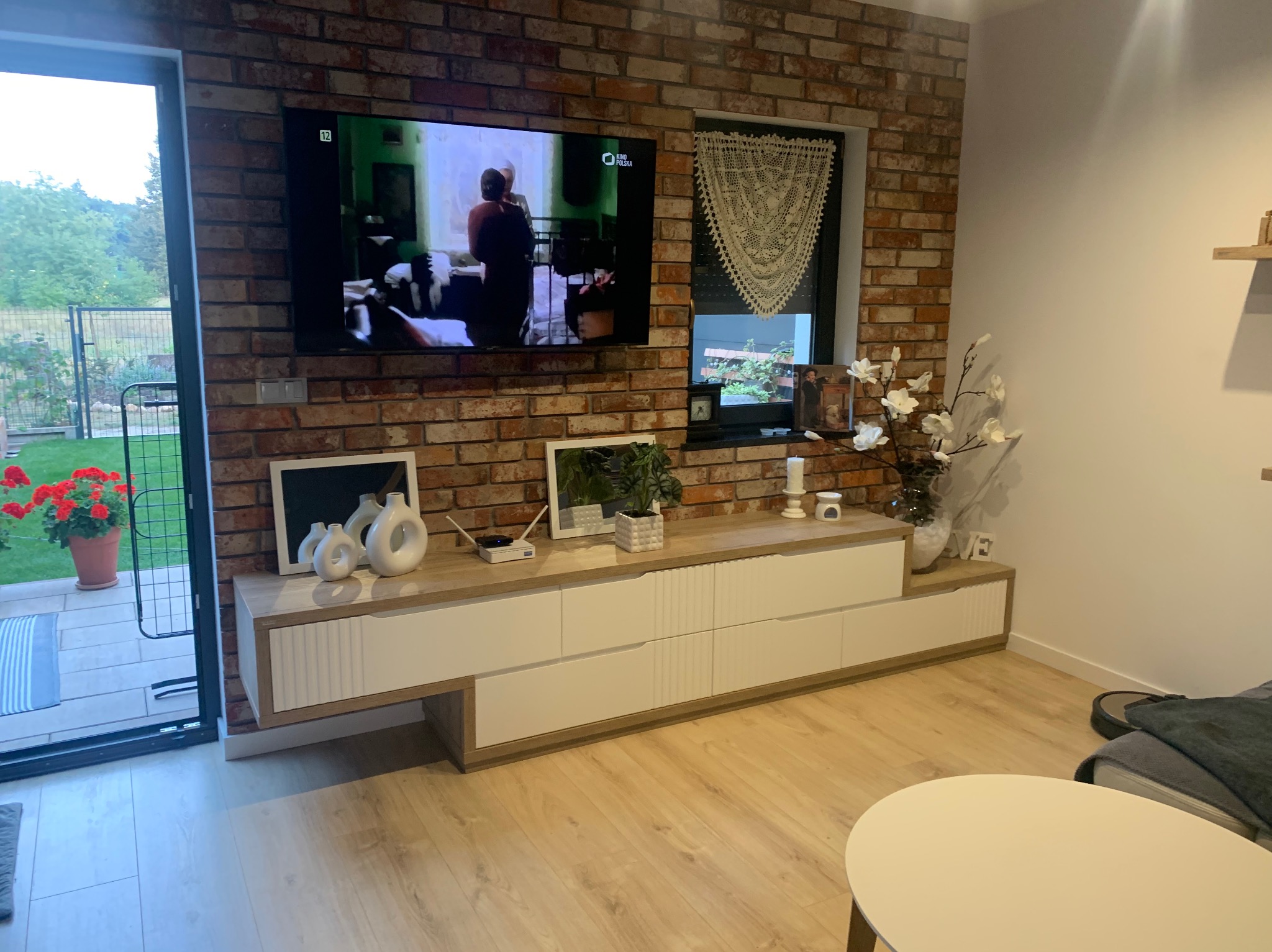 Salon z ceglaną ścianą, na której wisi telewizor i ozdobna serweta w oknie. Pod telewizorem biała szafka RTV z drewnianym blatem. Na blacie dekoracje, w tym białe ceramiczne wazony.