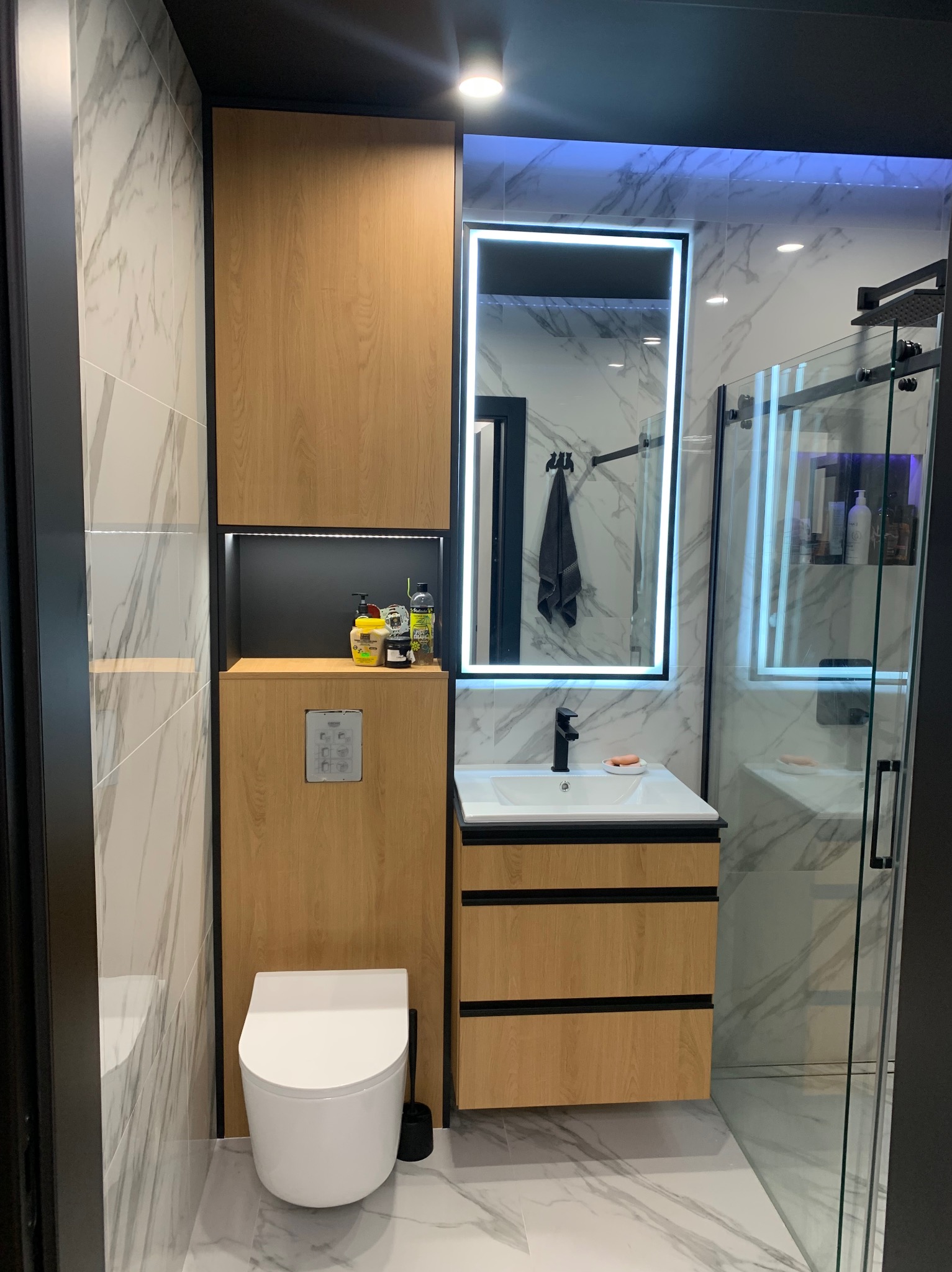 Nowoczesna łazienka z toaletą podwieszaną, szafką z umywalką i lustrem z podświetleniem LED, wykończona płytkami imitującymi marmur.