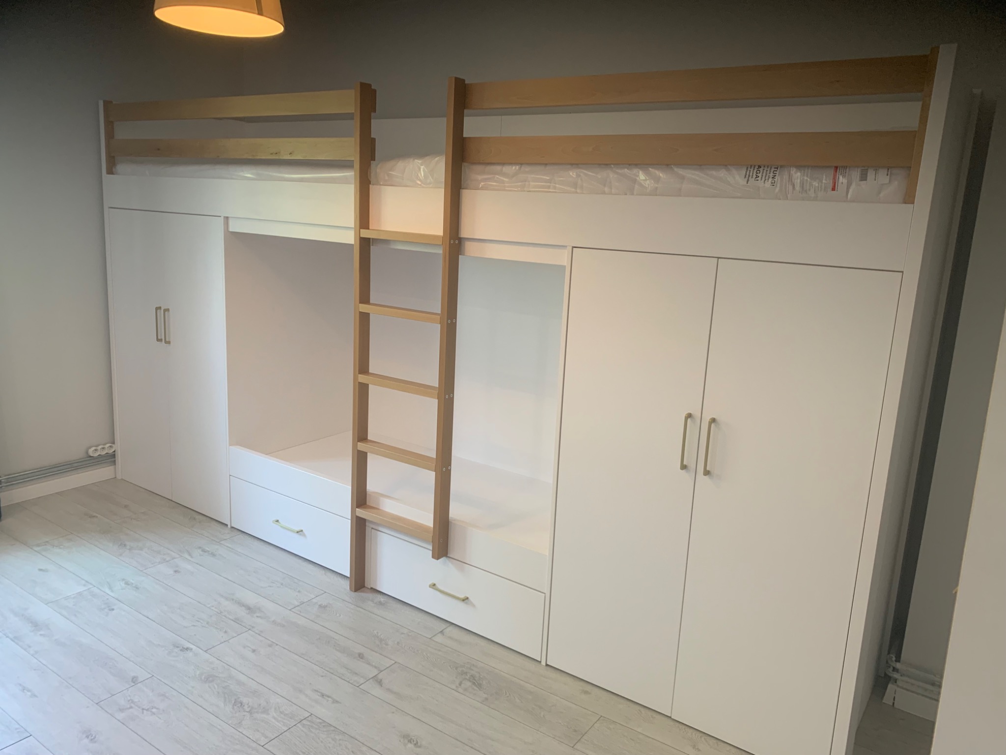 Biała antresola z drabinką i szafami po bokach, minimalistyczny design, drewniana drabinka, szare panele podłogowe, oświetlenie sufitowe.