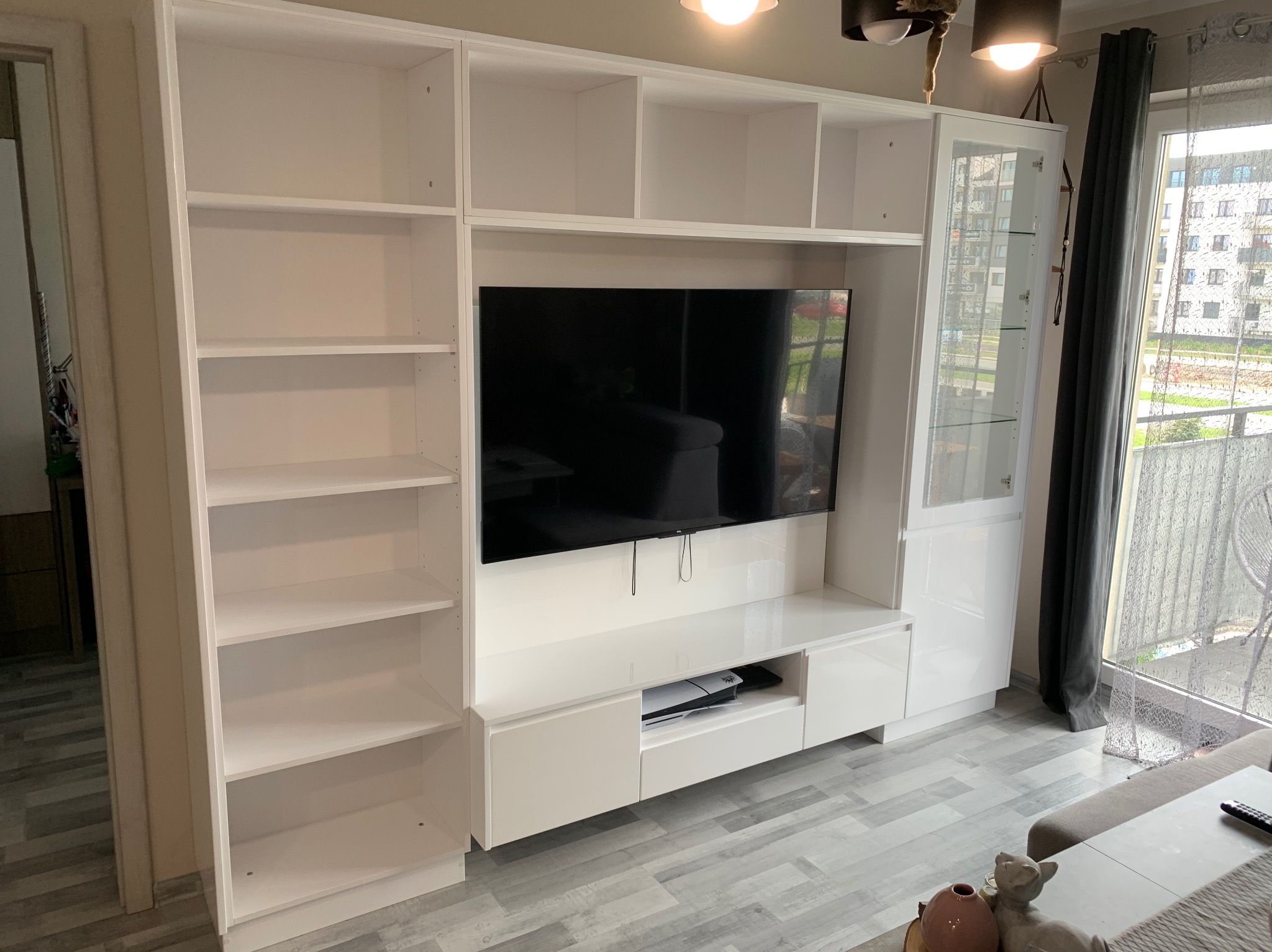 Biała szafa na wymiar z otwartymi półkami i szafkami pod telewizor, z widocznym dekoderem i konsolą do gier, ustawiona w salonie z szarymi zasłonami i widokiem na balkon.