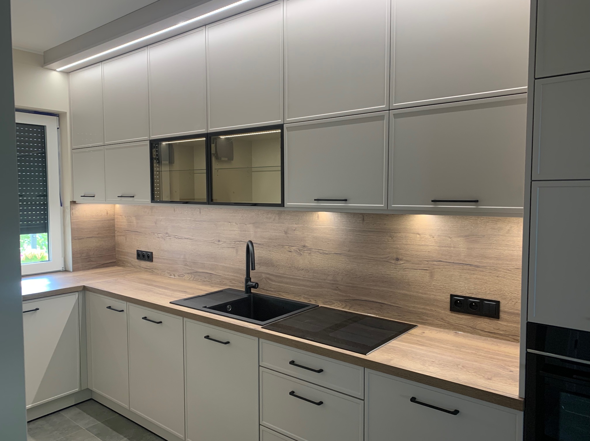 Nowoczesna kuchnia z białymi szafkami, drewnianym blatem i czarnym zlewem oraz płytą indukcyjną, oświetlenie LED pod szafkami górnymi.