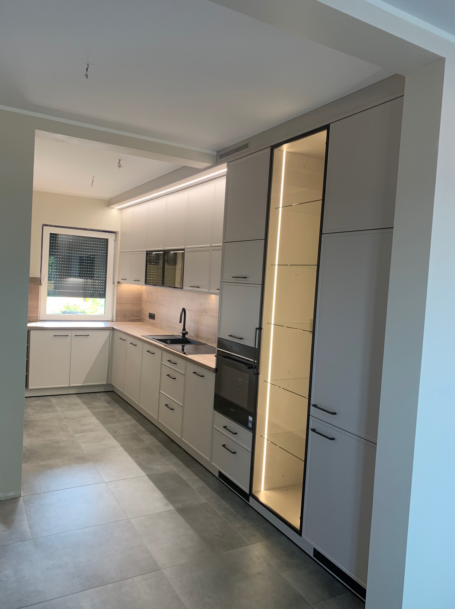 Nowoczesna kuchnia z szarymi frontami, czarnymi uchwytami i oświetleniem LED, widok na szafkę z przeszklonym frontem i podświetlonymi półkami.