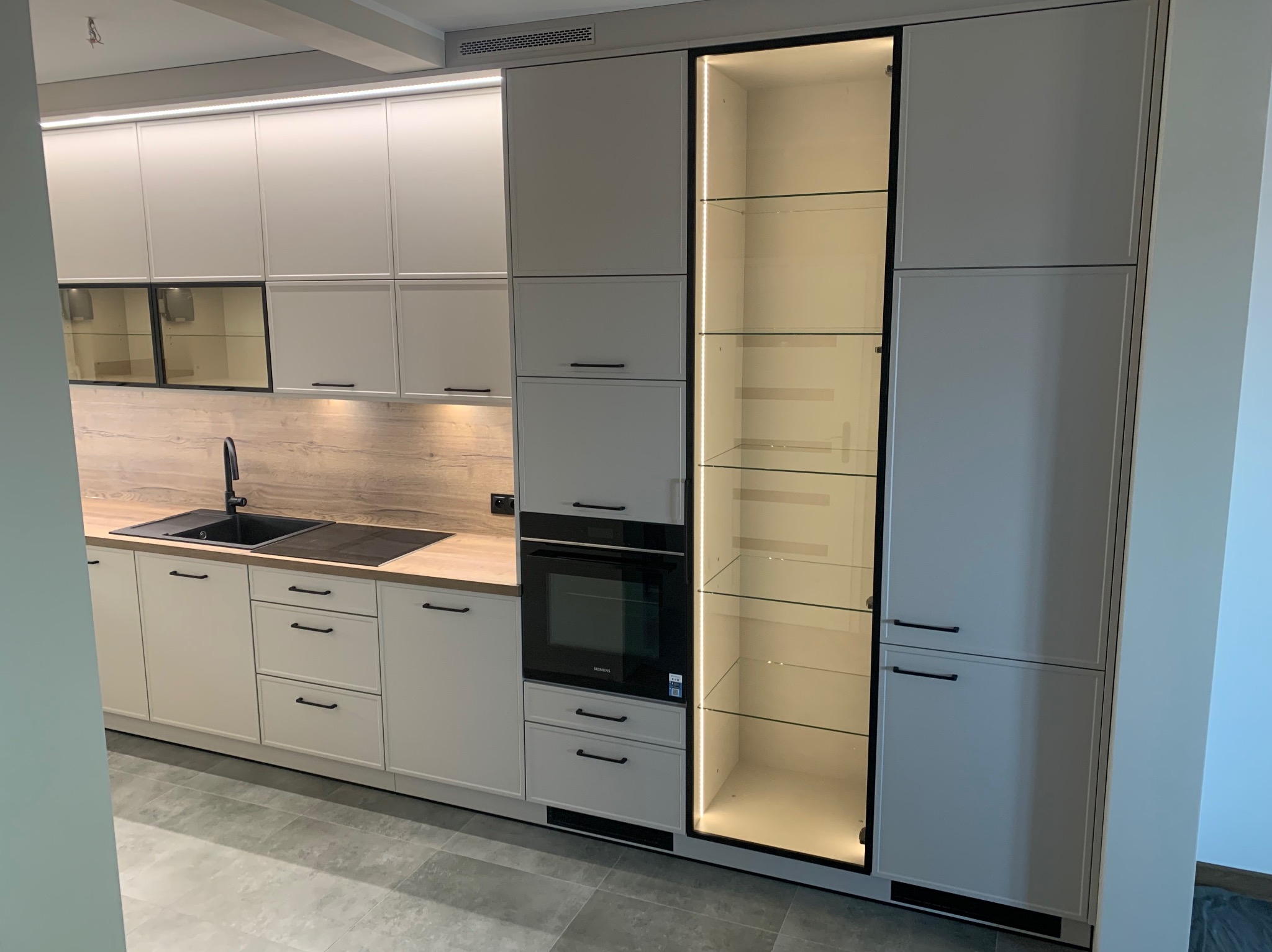 Jasna zabudowa kuchenna z beżowymi frontami, czarnymi uchwytami i ramami, drewnianym blatem oraz podświetlaną witryną z półkami ze szkła.