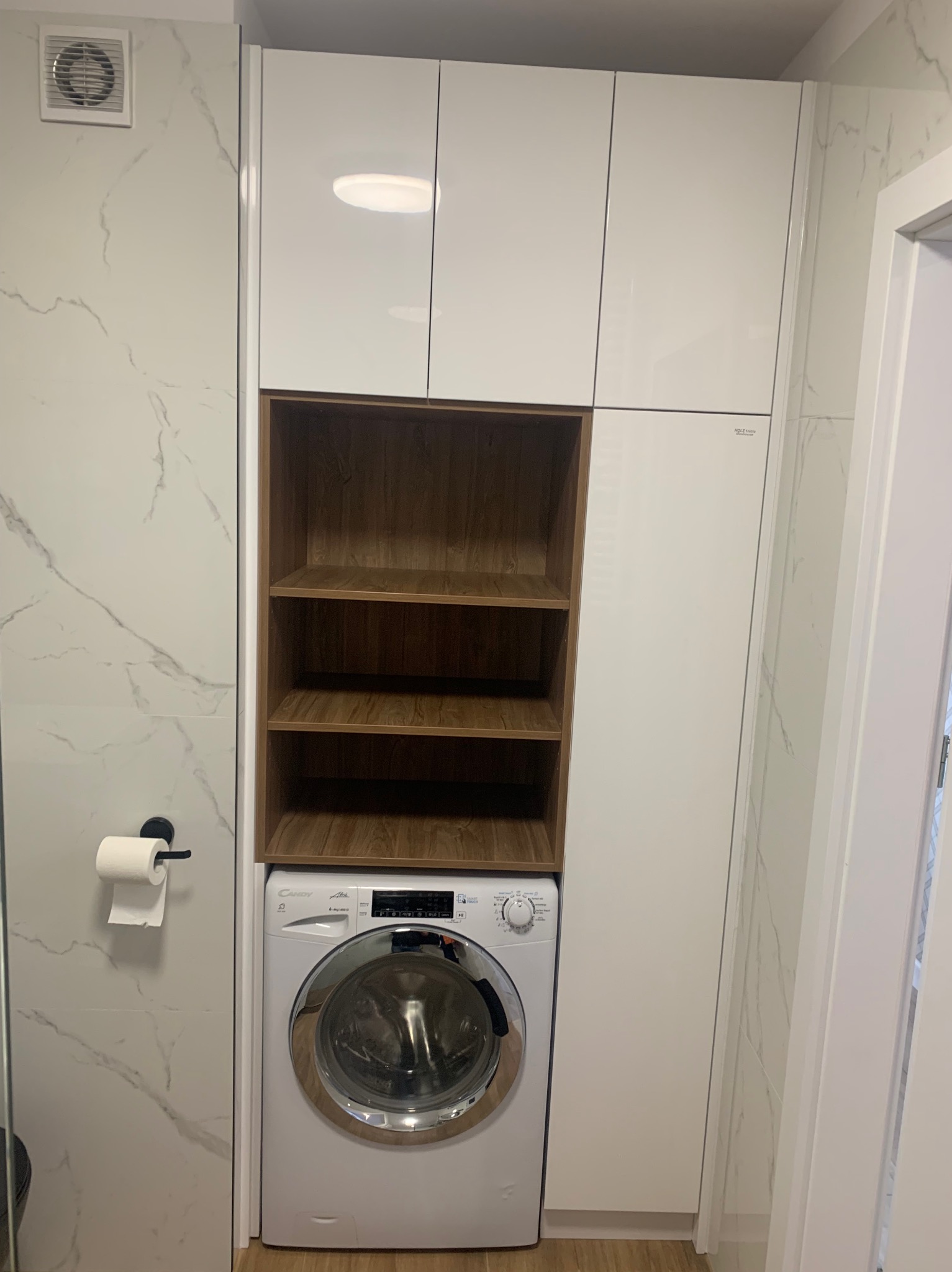 Biała zabudowa meblowa w łazience z pralką i drewnianymi półkami wnękowymi, widoczny fragment ściany z marmurowym wzorem i uchwyt na papier toaletowy.
