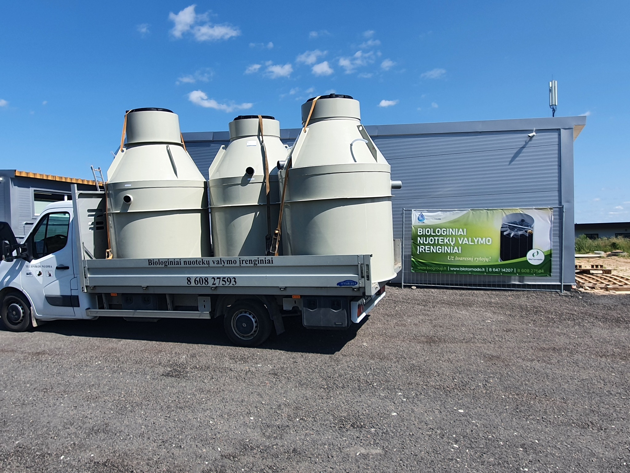 Trzy beżowe zbiorniki z drabinkami załadowane na samochód dostawczy, ustawione na żwirowym placu przed szarym budynkiem z anteną na dachu; w tle błękitne niebo.