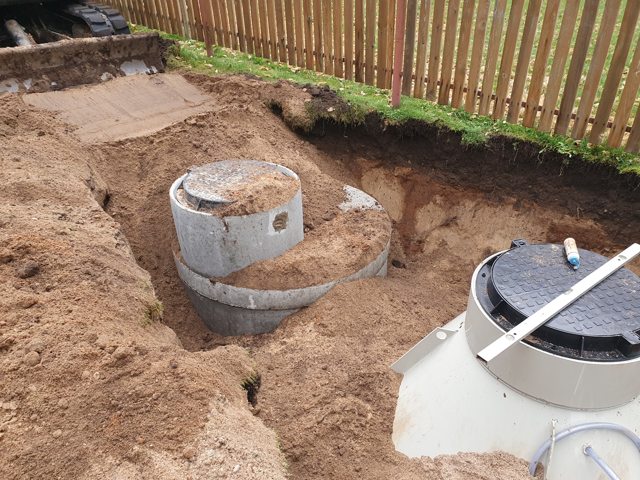 Betonowe kręgi szamba w wykopie, częściowo zasypane piaskiem, poziomica na pokrywie, w tle drewniany płot.