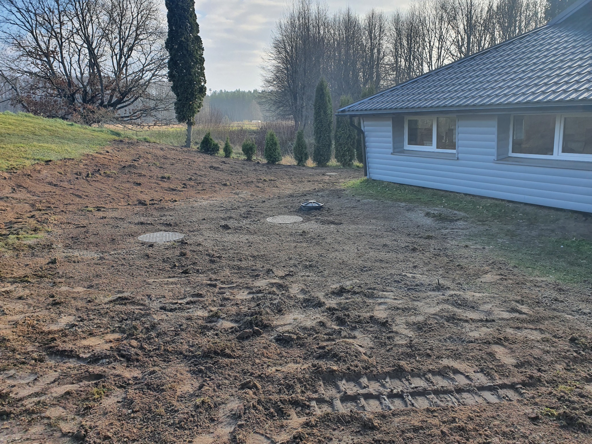 Teren po wykonanych pracach ziemnych z widocznymi włazami do podziemnych zbiorników, obok budynku mieszkalnego z nasadzeniami iglaków.