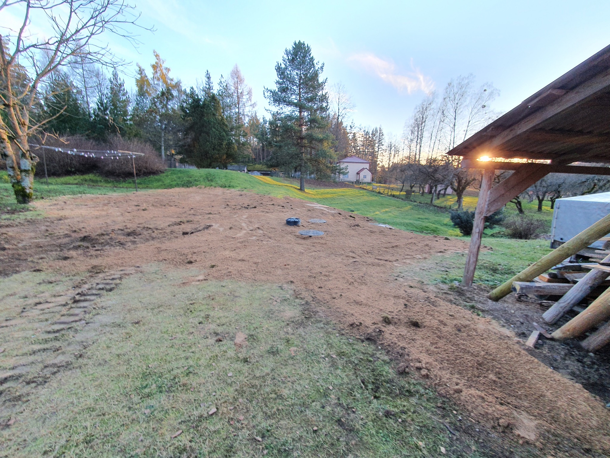 Przygotowany teren pod budowę, prawdopodobnie pod instalację sanitarną, z widocznymi śladami opon i pokrywą włazu w tle, na tle zieleni i zabudowań mieszkalnych w oddali.