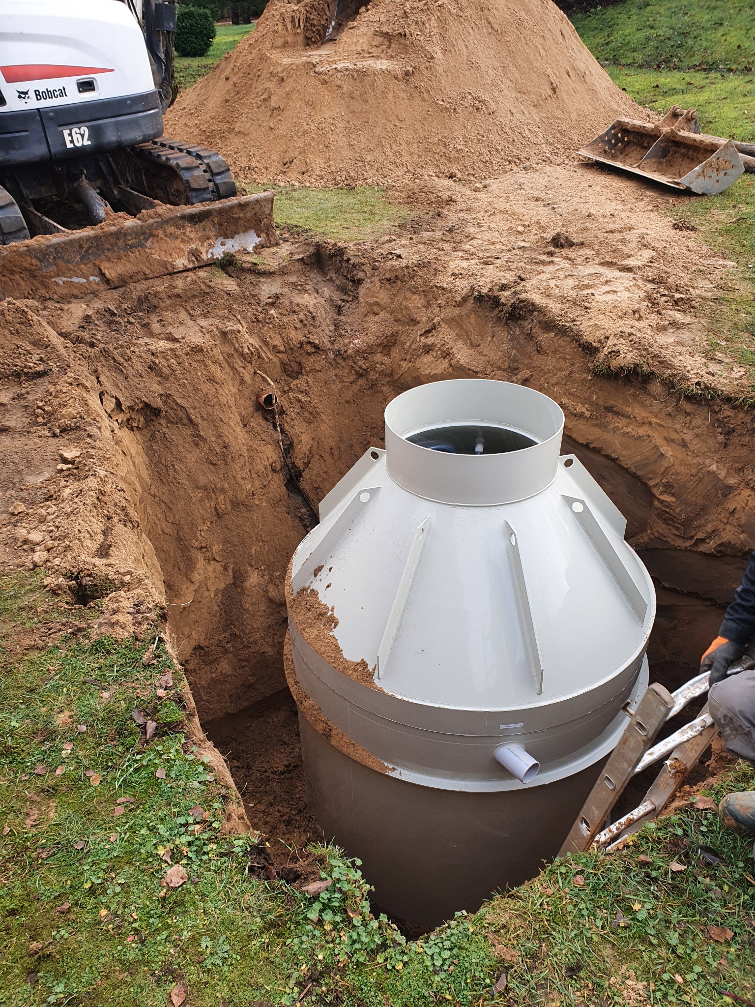 Montaż szarej, plastikowej konstrukcji zbiornika w wykopie ziemnym; w tle koparka Bobcat i pryzma piasku.