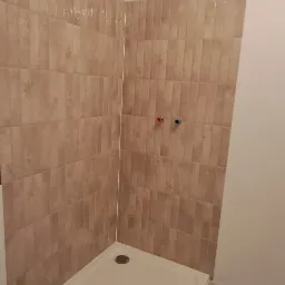 Wykończona kabina prysznicowa z beżowymi płytkami ułożonymi pionowo, widoczne dwa zawory z oznaczeniem ciepłej i zimnej wody, oraz odpływ w brodziku.