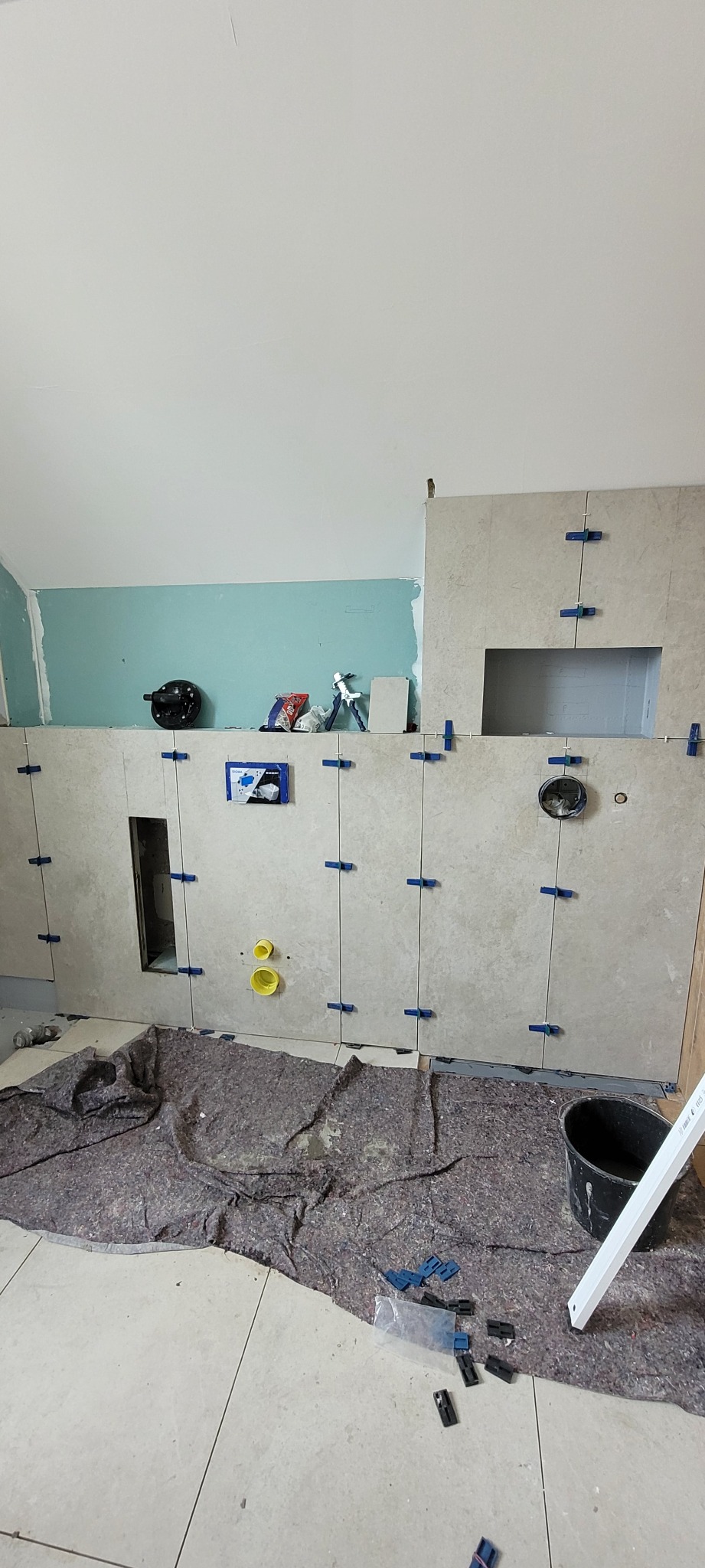 Ściana w trakcie układania płytek ceramicznych z widocznymi krzyżykami dystansowymi, przygotowane otwory na instalacje, zabezpieczona podłoga i narzędzia pracy.