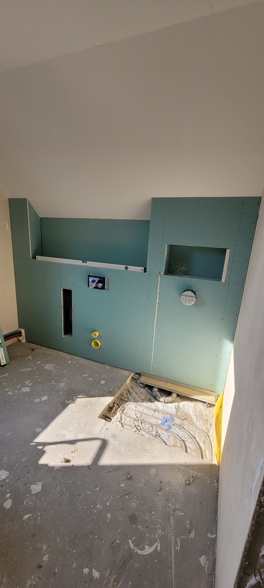 Ściana z płyty karton gips, widoczne otwory na instalacje, poziomica, żółte rury, fragment ogrzewania podłogowego i surowy beton na podłodze.