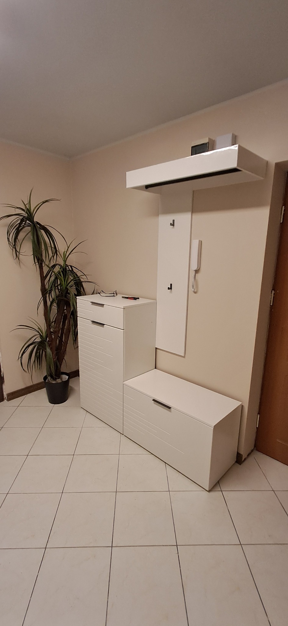 Nowoczesny zestaw mebli do przedpokoju w kolorze białym, składający się z szafki na buty z siedziskiem, komody z szufladami oraz panelu z wieszakami i półką, uzupełniony o doniczkową palmę.
