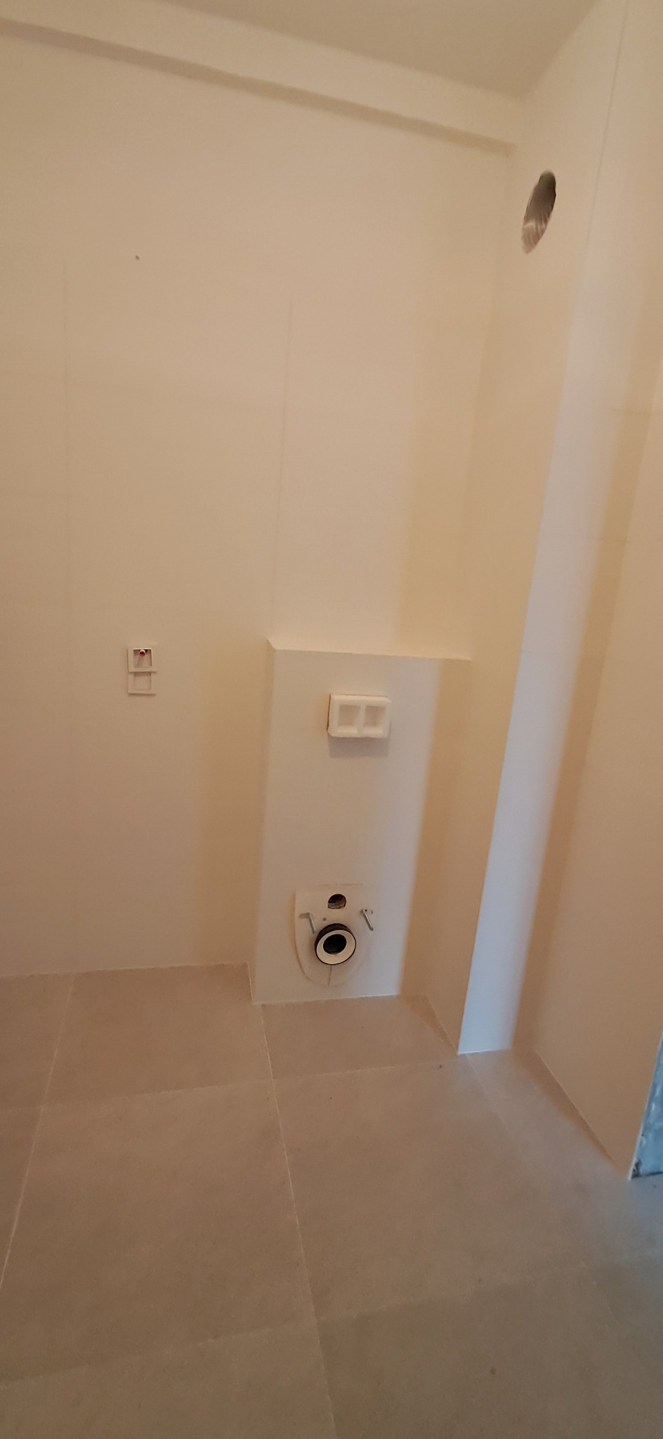 Surowe wnętrze łazienki w trakcie remontu, widoczne płytki podłogowe, instalacja podtynkowego WC, otwór w ścianie i puszki elektryczne.