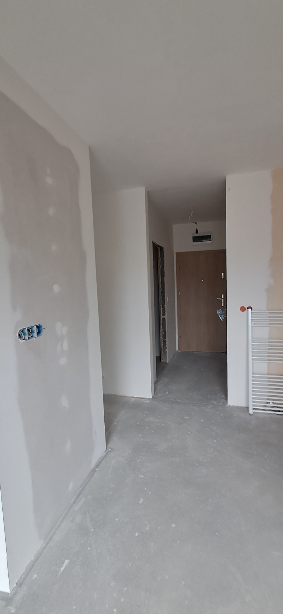 Wnętrze w trakcie prac wykończeniowych: ściany po szpachlowaniu, widoczne puszki elektryczne, surowa podłoga, grzejnik drabinkowy w tle.