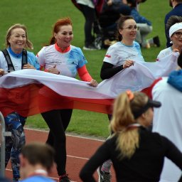 Artur Pawlak - Grupa uśmiechniętych kobiet biegnie po bieżni trzymając biało-czerwoną flagę, w tle zielona trawa i widzowie.