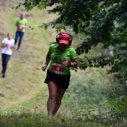 Artur Pawlak - Kobieta w czerwonej czapce i słuchawkach biegnie pod górę podczas biegu przełajowego z numerem 4 na koszulce, w tle dwie inne osoby.