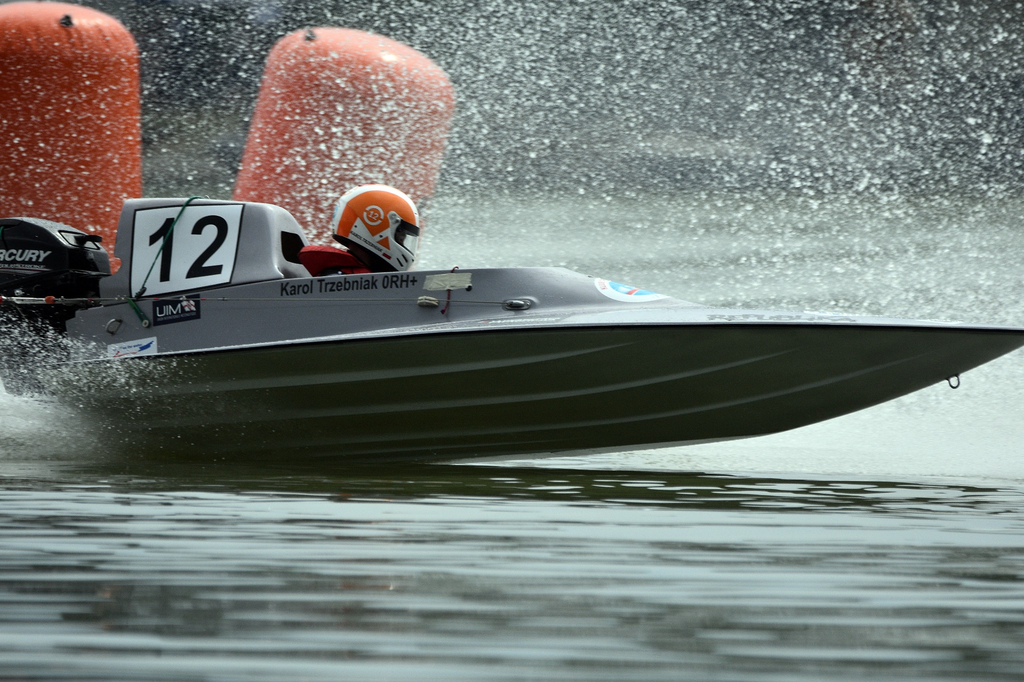 Szara motorówka z numerem 12, pilot w pomarańczowym kasku, pędzi po wodzie, za nią rozprysk wody i pomarańczowe boje.