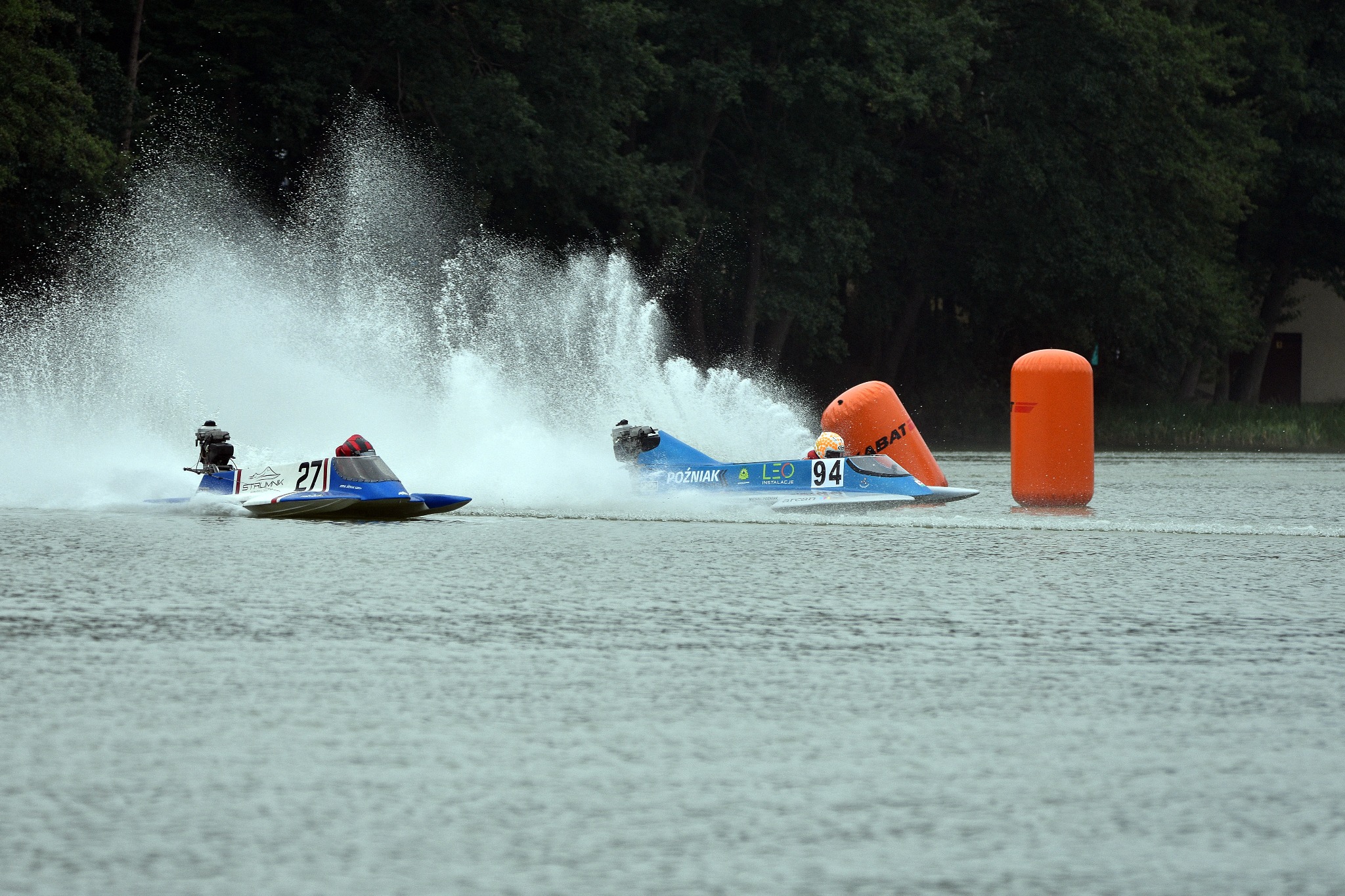 Dwie motorówki ścigają się na wodzie, tworząc fontanny wody, z widocznymi numerami 271 i 94, oraz pomarańczowymi bojami wyznaczającymi tor.