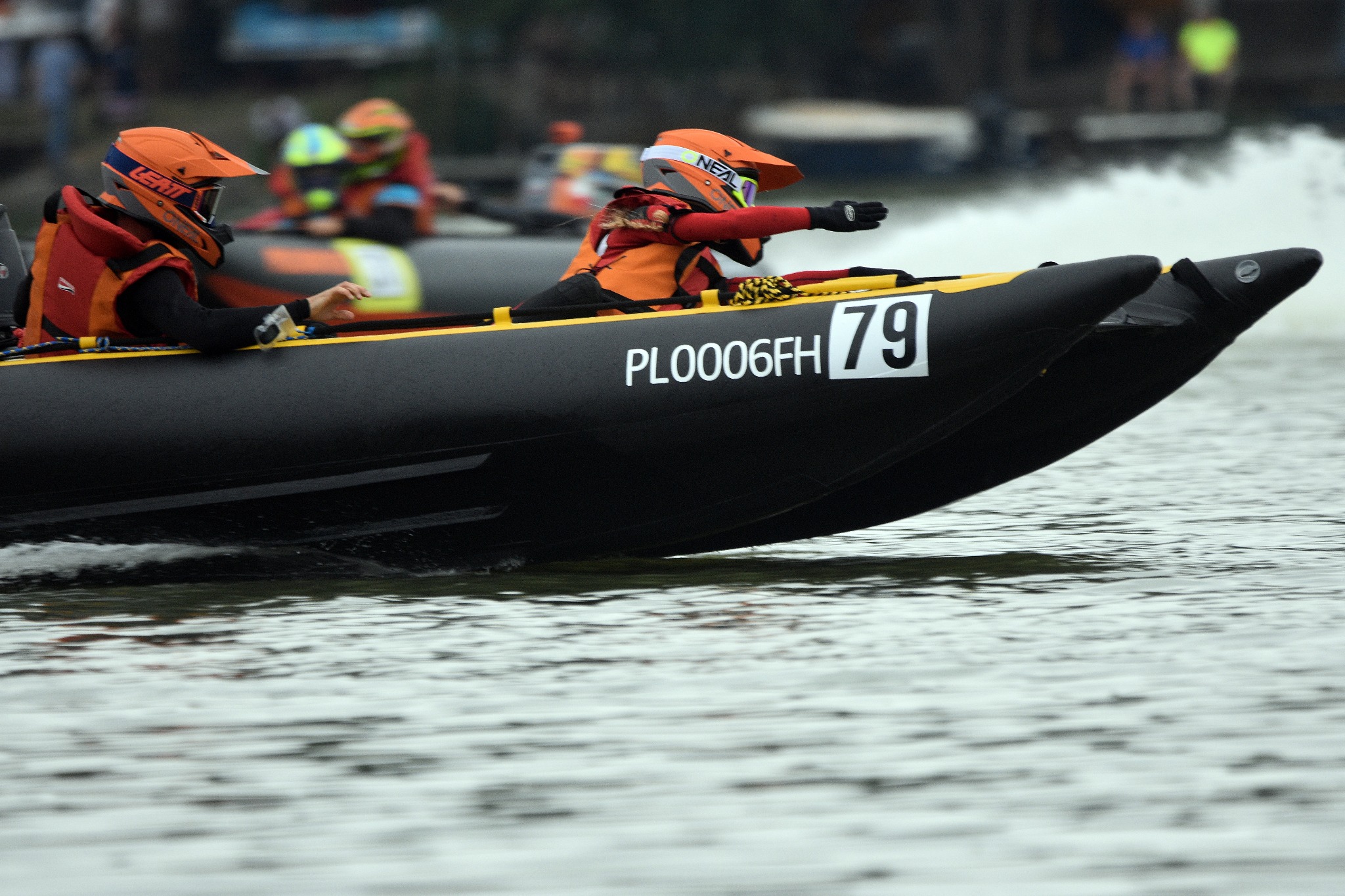 Dwie osoby w jaskrawo pomarańczowych kaskach i kamizelkach ratunkowych ścigają się na czarnej łodzi motorowej z numerem 79 i rejestracją PL0006FH po wzburzonej wodzie.