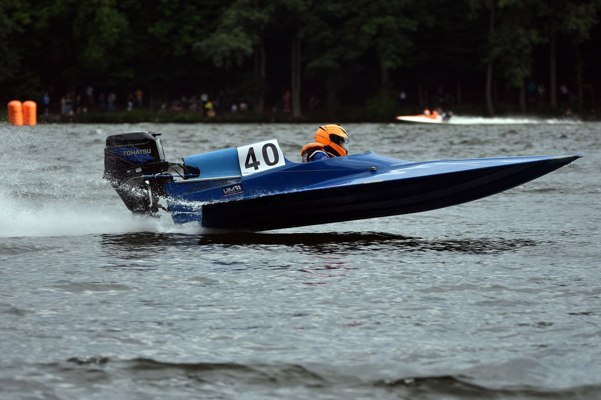 Niebieska łódź motorowa z numerem 40 w pełnym pędzie po wodzie, z pomarańczowym kaskiem kierowcy i silnikiem Tohatsu, w tle zalesiony brzeg.