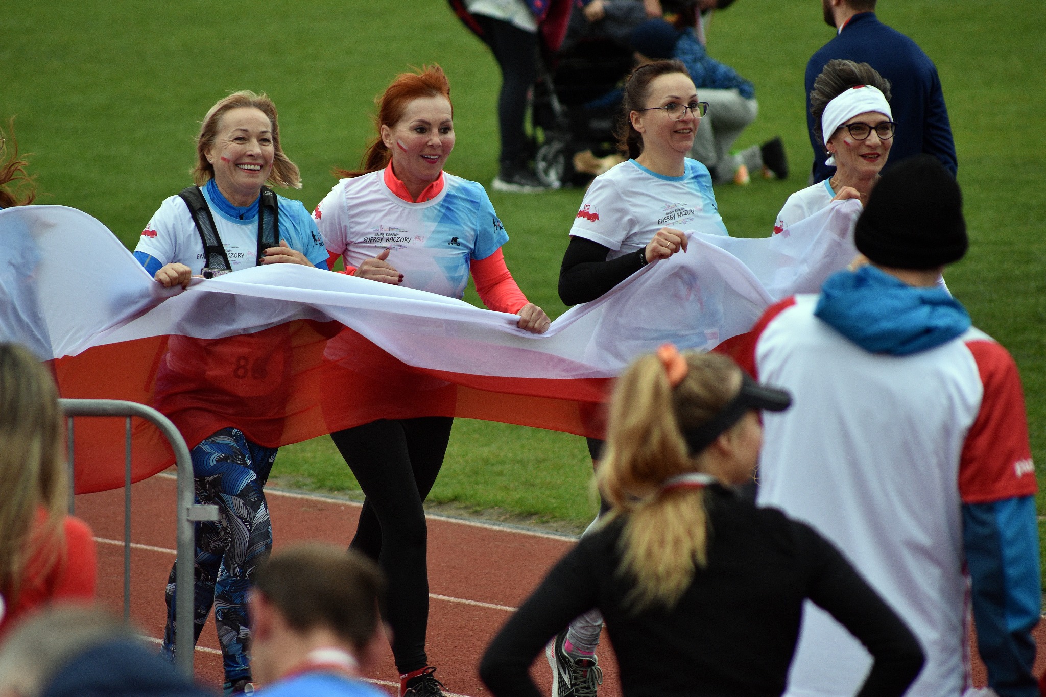 Grupa uśmiechniętych kobiet biegnie po bieżni trzymając biało-czerwoną flagę, w tle zielona trawa i widzowie.