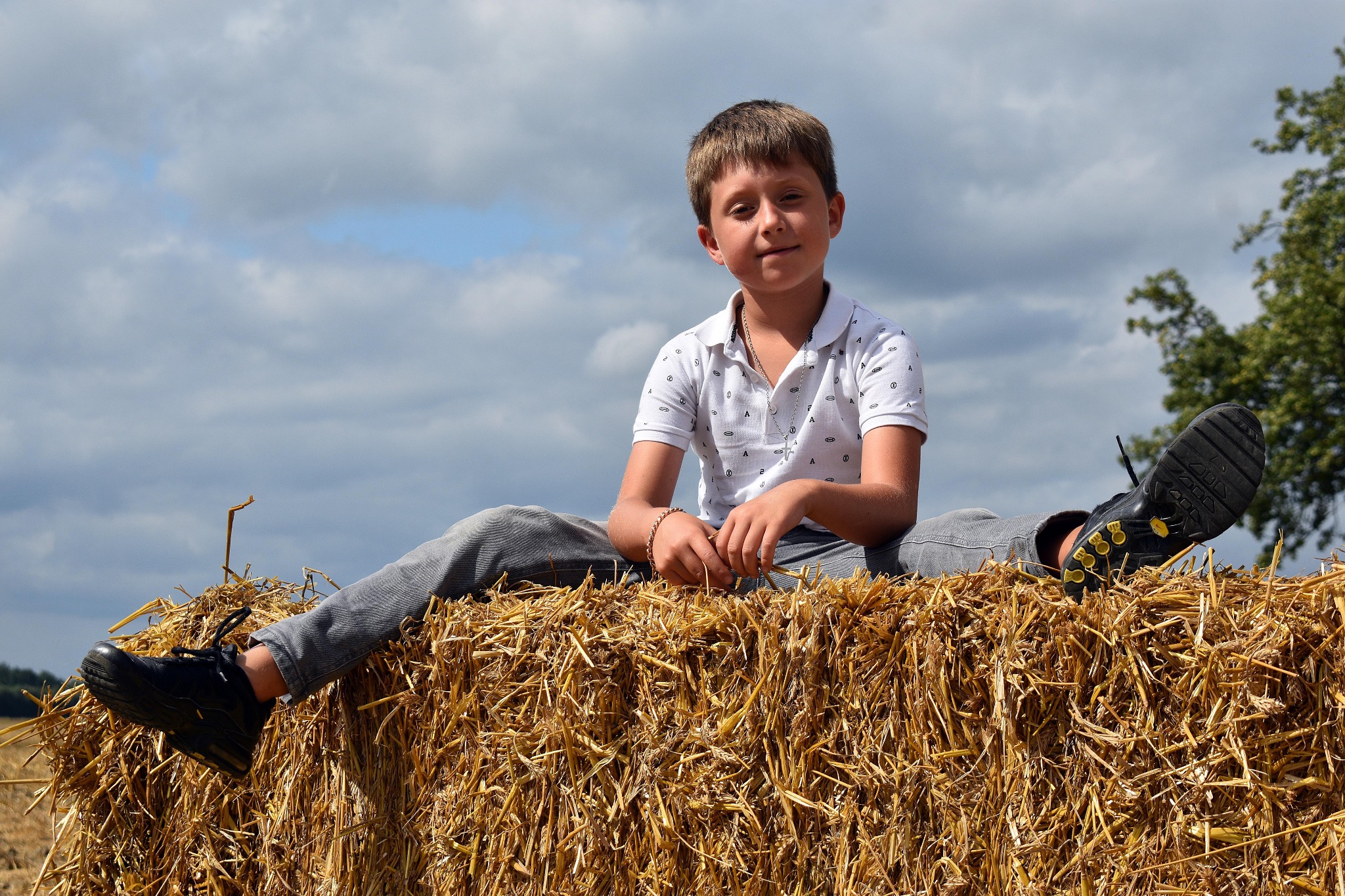 Chłopiec w białej koszulce polo i szarych spodniach siedzi na belocie słomy na polu, z nogami wyciągniętymi na boki, na tle pochmurnego nieba.