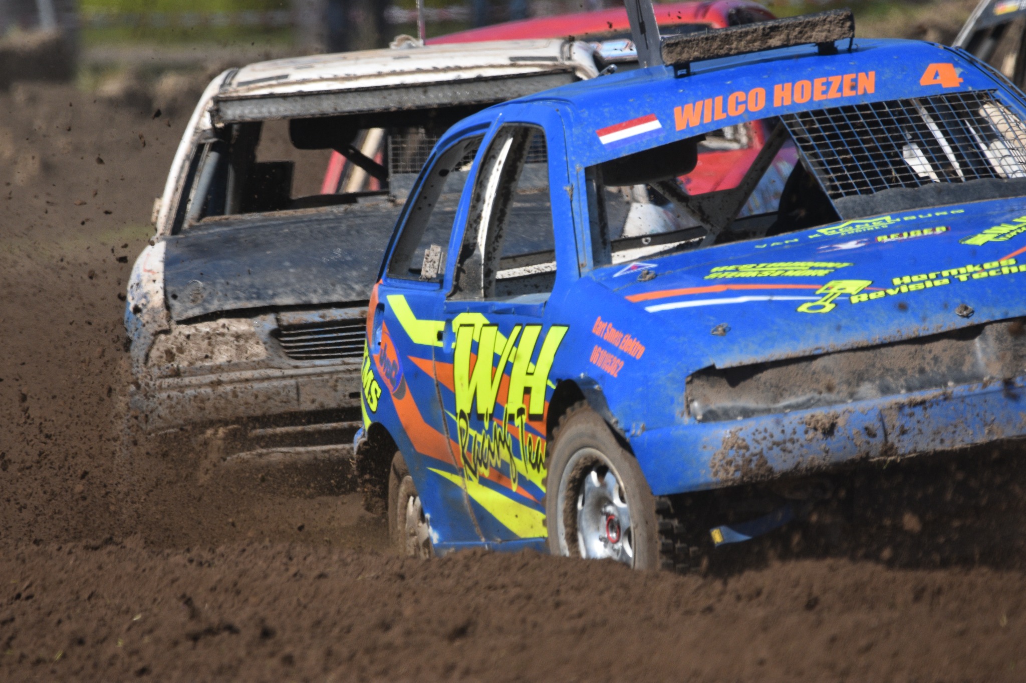 Dwa samochody biorące udział w wyścigu demolition derby, pokryte błotem i uszkodzeniami, w trakcie dynamicznej akcji na torze.