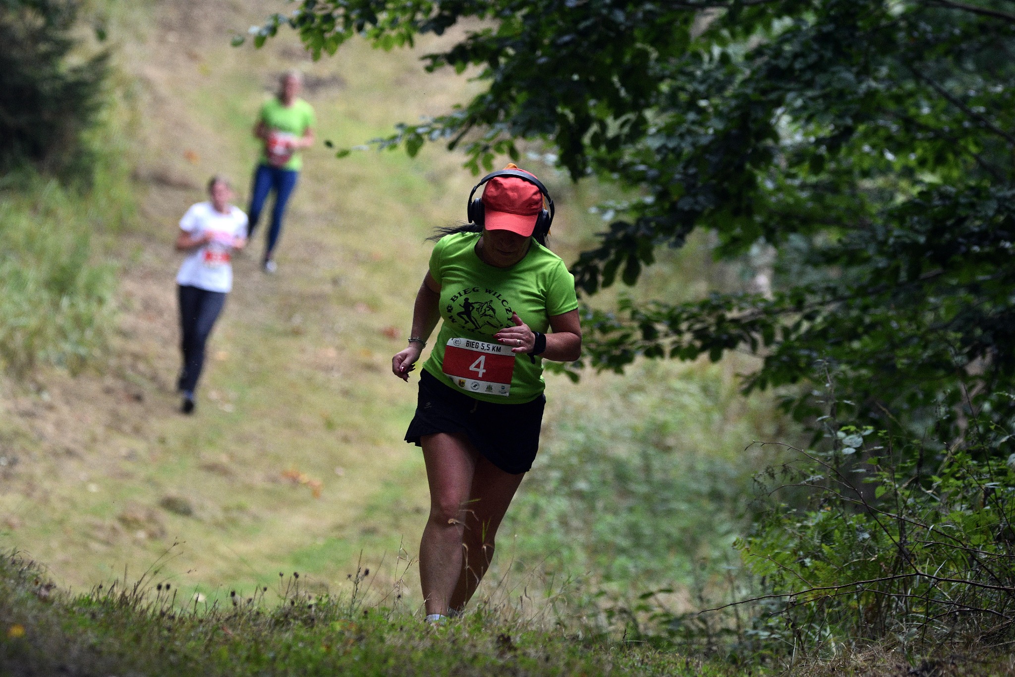 Kobieta w czerwonej czapce i słuchawkach biegnie pod górę podczas biegu przełajowego z numerem 4 na koszulce, w tle dwie inne osoby.