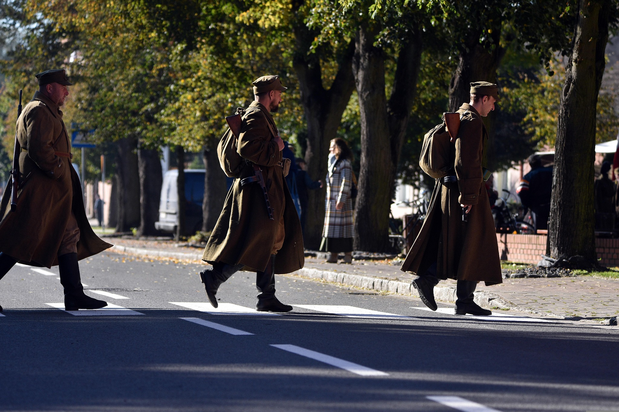 Trzech mężczyzn w historycznych, wojskowych uniformach z karabinami przechodzi przez przejście dla pieszych na tle drzew i budynków.
