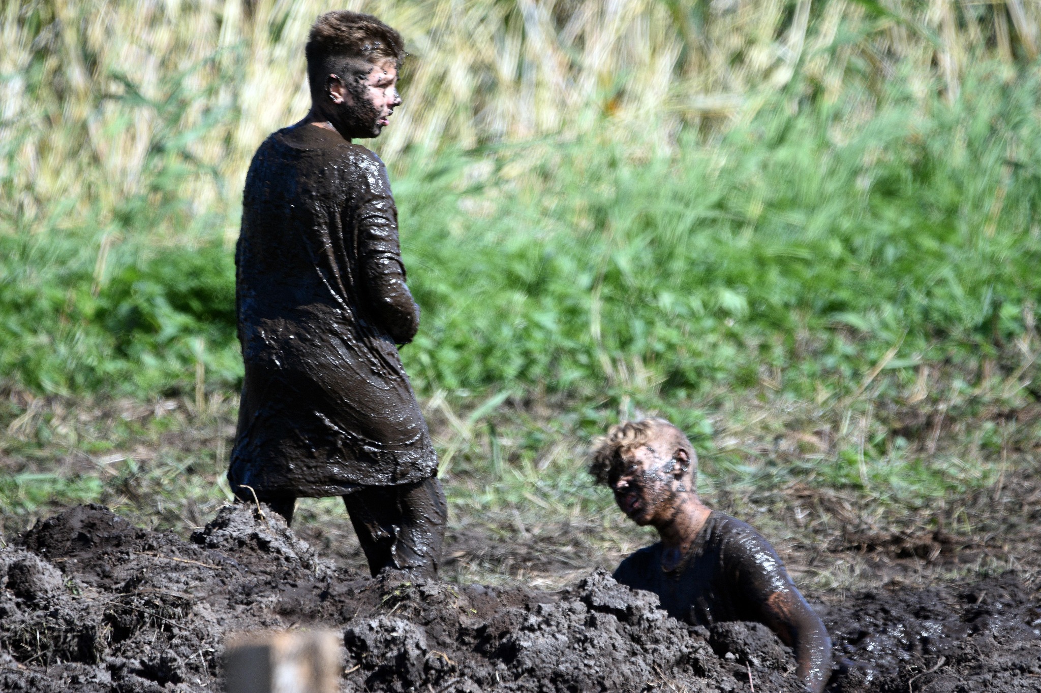 Dwaj mężczyźni cali w błocie podczas ekstremalnego biegu z przeszkodami, jeden stoi, drugi zanurzony w błocie po pas, w tle trawa i zarośla.