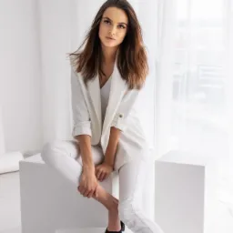 Kobieta w białym garniturze pozuje siedząc na białych kostkach na tle białych zasłon, czarne szpilki, naturalne światło.