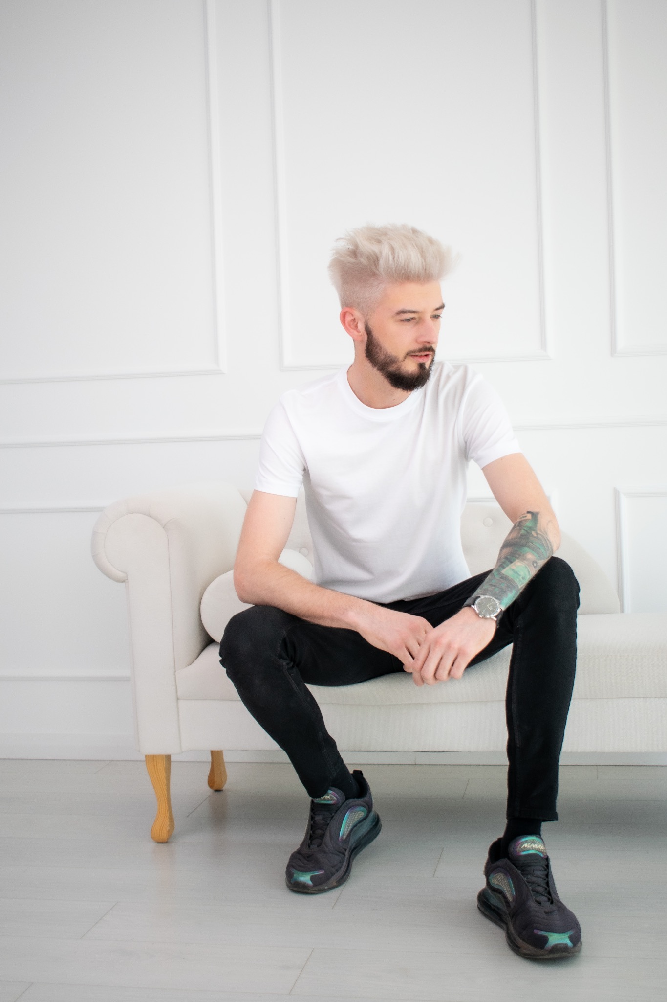 Młody mężczyzna z platynowymi włosami i tatuażem na ramieniu siedzi na białej sofie, ubrany w biały t-shirt i czarne jeansy, spoglądając w bok.