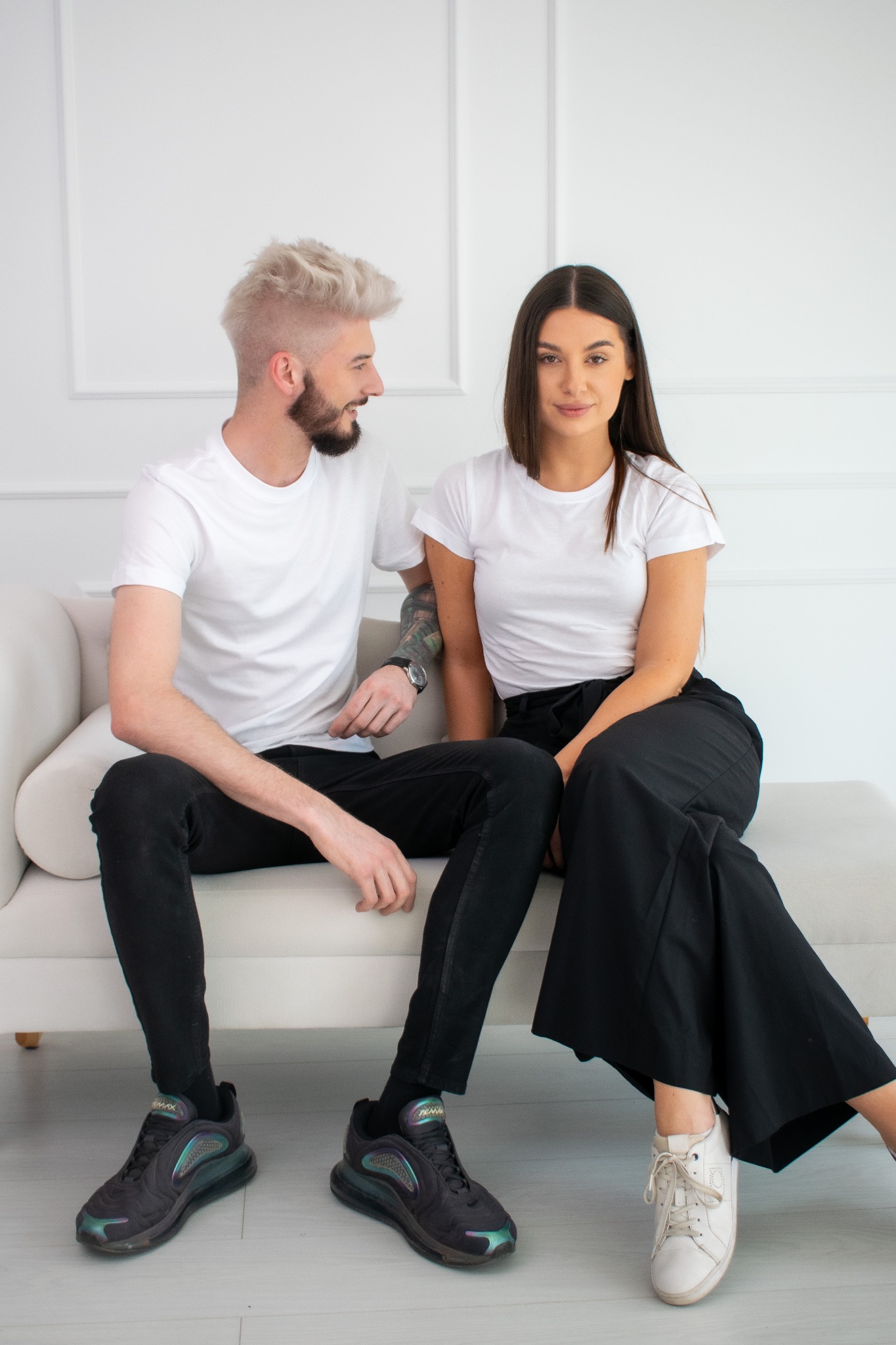 Para siedząca na białej sofie: mężczyzna z platynowymi włosami i tatuażem na ramieniu, kobieta w czarnych spodniach i białych trampkach.