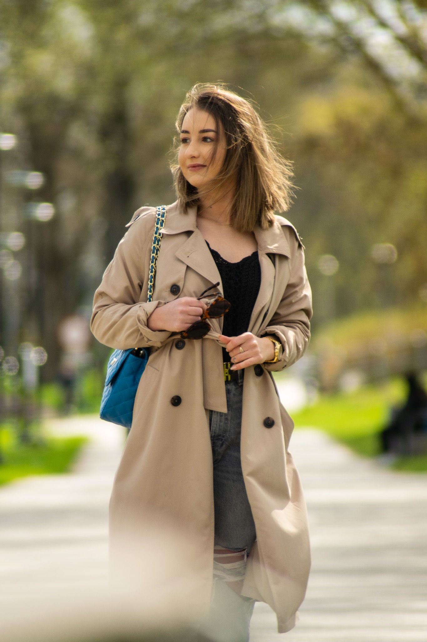 Kobieta w beżowym trenczu, z niebieską torebką na łańcuszku, trzymająca okulary przeciwsłoneczne, spaceruje alejką parkową w słoneczny dzień.