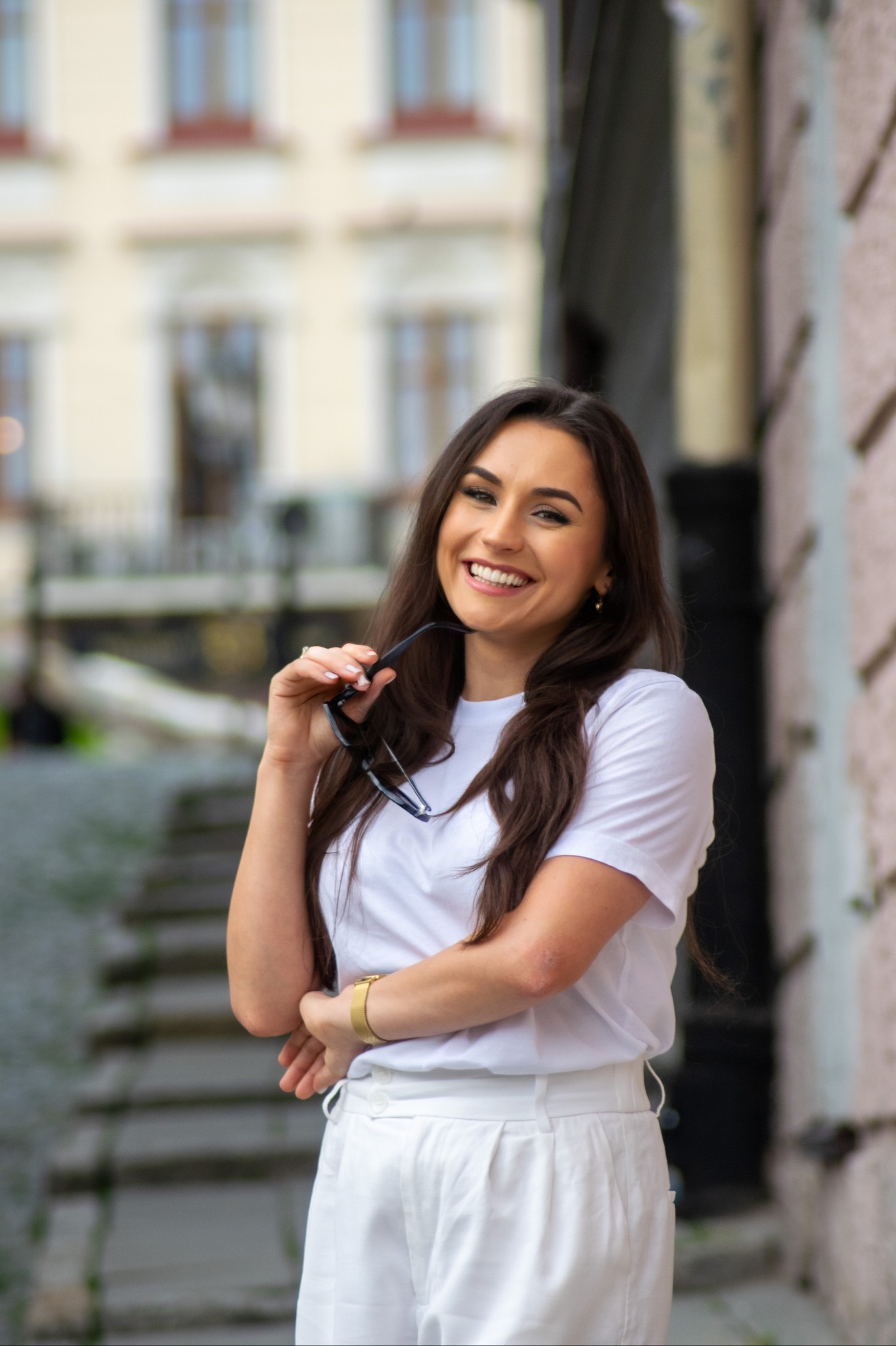 Uśmiechnięta brunetka w białym stroju, trzymająca okulary w dłoni, pozuje na tle miejskiej architektury z widocznymi schodami.
